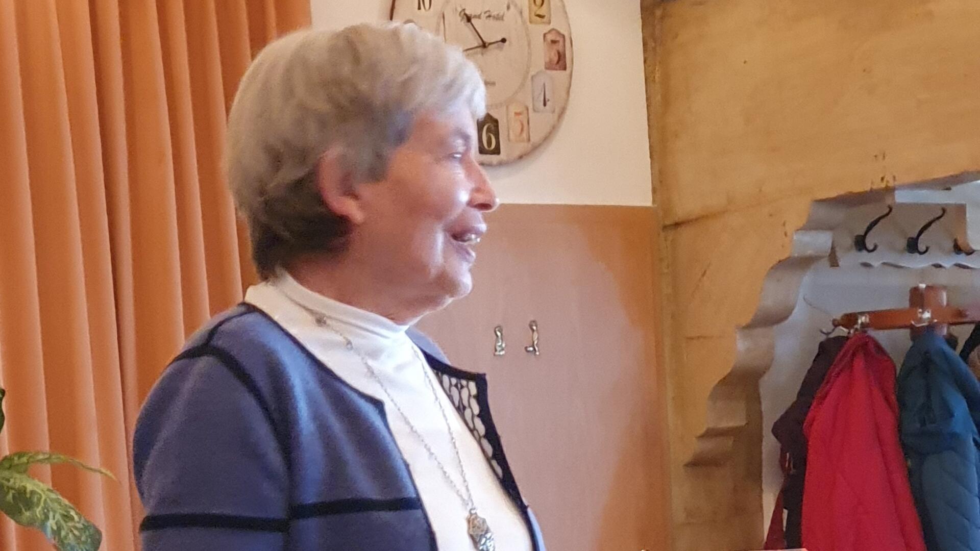 Eine ältere Frau mit grauen Haaren und blau-weißem Outfit spricht lächelnd vor einer Gruppe.