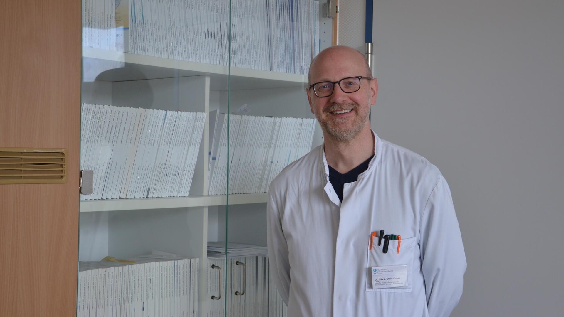 Ein Arzt steht in weißem Kittel vor einem Bücherregal und lächelt in die Kamera.