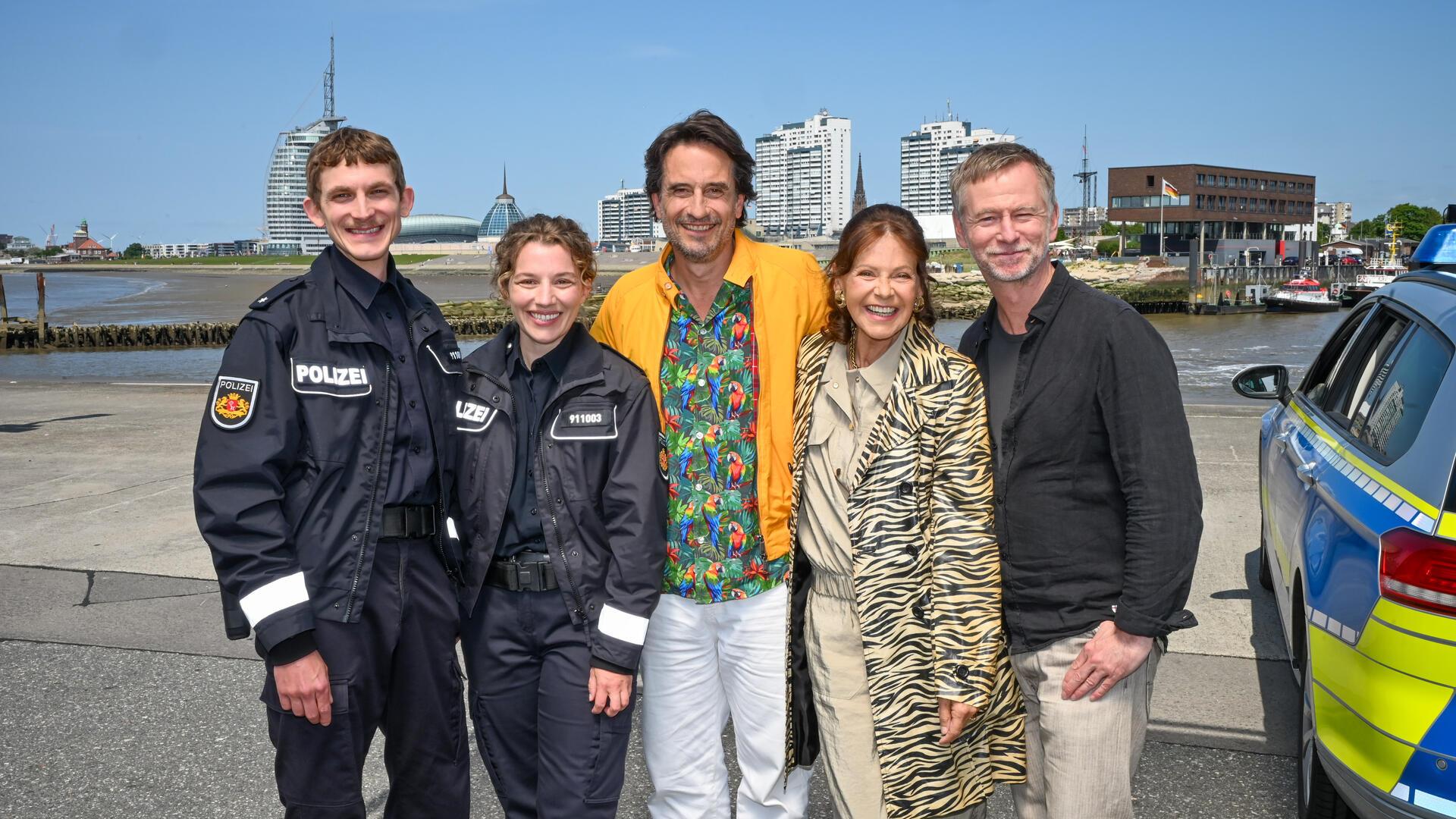 Fünf Personen lächeln vor der Skyline einer Stadt in die Kamera. Rechts an der Seite steht ein Polizeiauto.