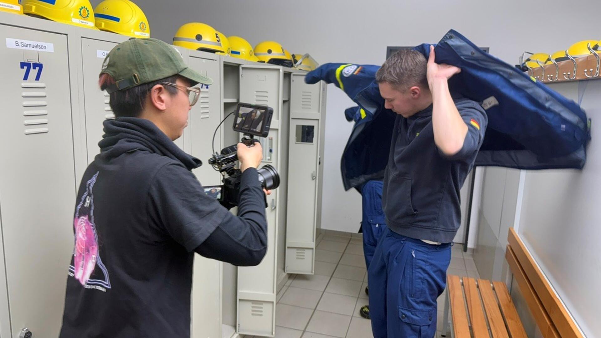  In einer Umkleide eines Hilfswerks zieht sich ein junger Mann eine blaue Einsatzjacke über, während ein Kameramann mit Kappe und schwarzem T-Shirt ihn mit einer professionellen Kamera filmt. Im Hintergrund sind Spinde mit gelben Schutzhelmen zu sehen.