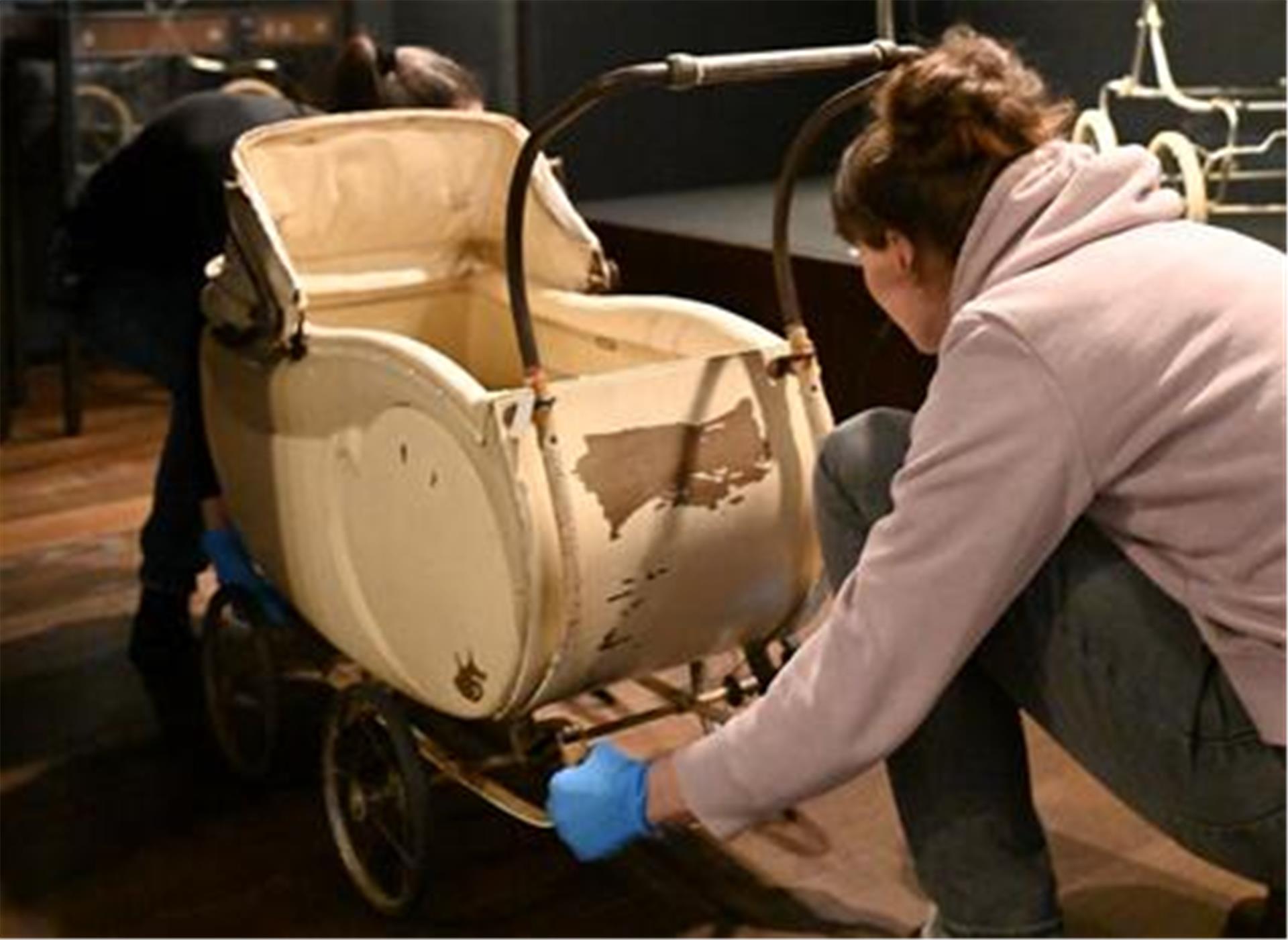 Eine Frau mit blauen Handschuhen schiebt einen alten, beigen Kinderwagen mit abgenutzter Lackierung in eine Museumsausstellung.