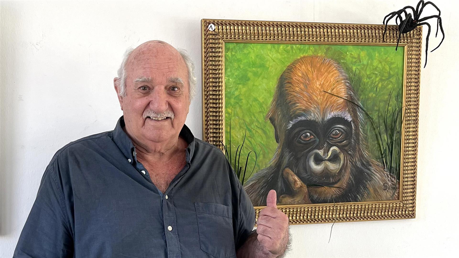 Ein älterer Mann mit schütterem grauen Haar und einem grauen Oberlippenbart steht vor einem Kunstwerk und zeigt mit seiner linken Hand auf das Gemälde - auf dem ist ein Affe (Gorilla?) zu sehen. Am oberen Rand des Gemäldes scheint eine schwarze Spinne (schwarze Witwe?) zu sitzen.