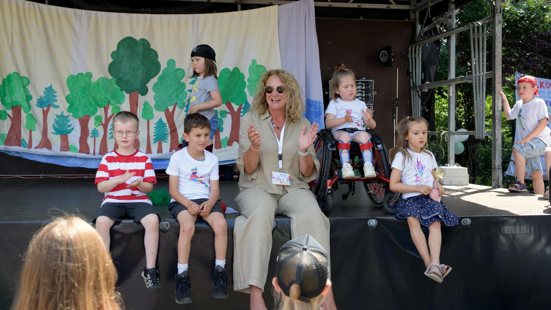 Eine Gruppe von Kindern und eine Erzieherin sitzen auf einer Bühne vor einem gemalten Waldhintergrund. Ein Mädchen im Rollstuhl ist ebenfalls Teil der Gruppe. Alle Beteiligten wirken fröhlich und entspannt bei sommerlichem Wetter.