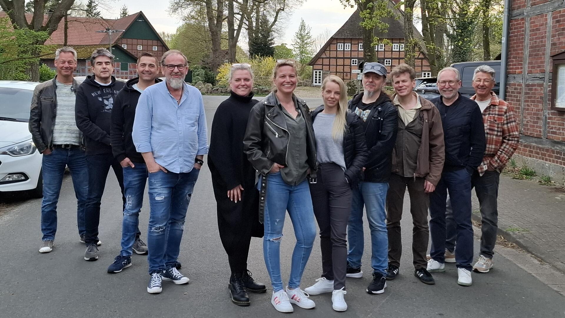 Gruppe von elf Personen, darunter Männer und Frauen, steht lächelnd auf einer Dorfstraße vor Fachwerkhäusern; sie tragen Freizeitkleidung und wirken gut gelaunt.
