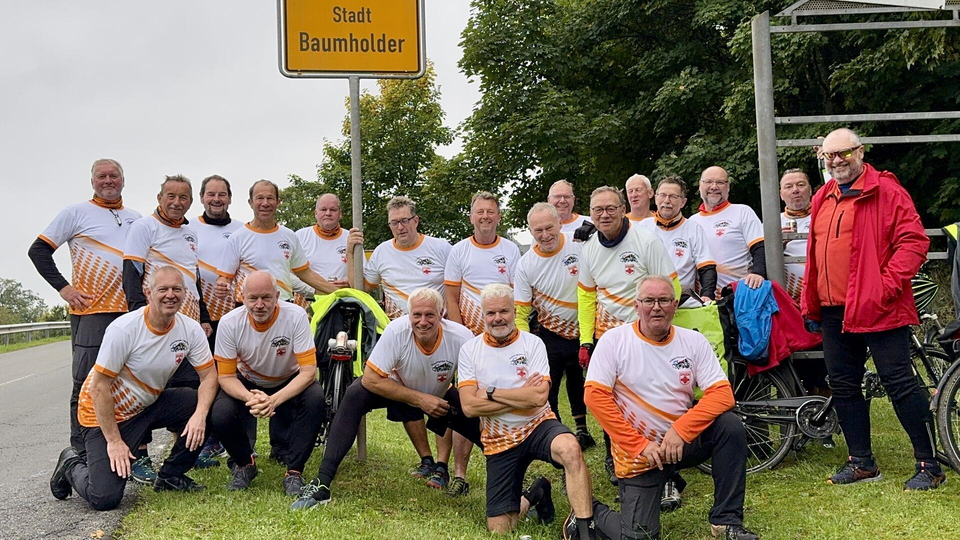 Eine Gruppe von 19 Radfahrern in einheitlichen Trikots steht lächelnd unter dem Ortsschild der Stadt Baumholder. Im Hintergrund sind Bäume und mehrere Fahrräder zu sehen.