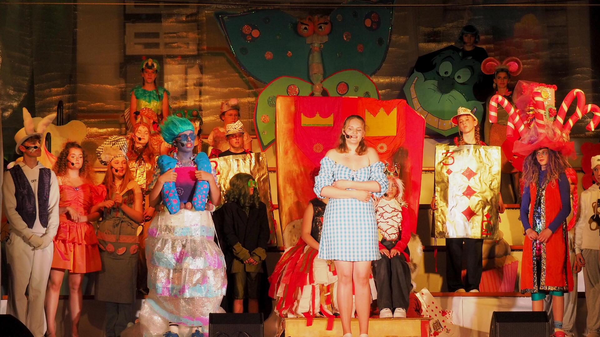 Schülerinnen und Schüler des Gymnasiums Wesermünde in bunten Kostümen auf der Bühne während des Musicals „Alice“. Im Mittelpunkt steht eine Schülerin im blau-weißen Kleid als Alice, umgeben von fantasievoll verkleideten Figuren wie Karten, Tieren und Fabelwesen vor einer farbenfrohen Kulisse.
