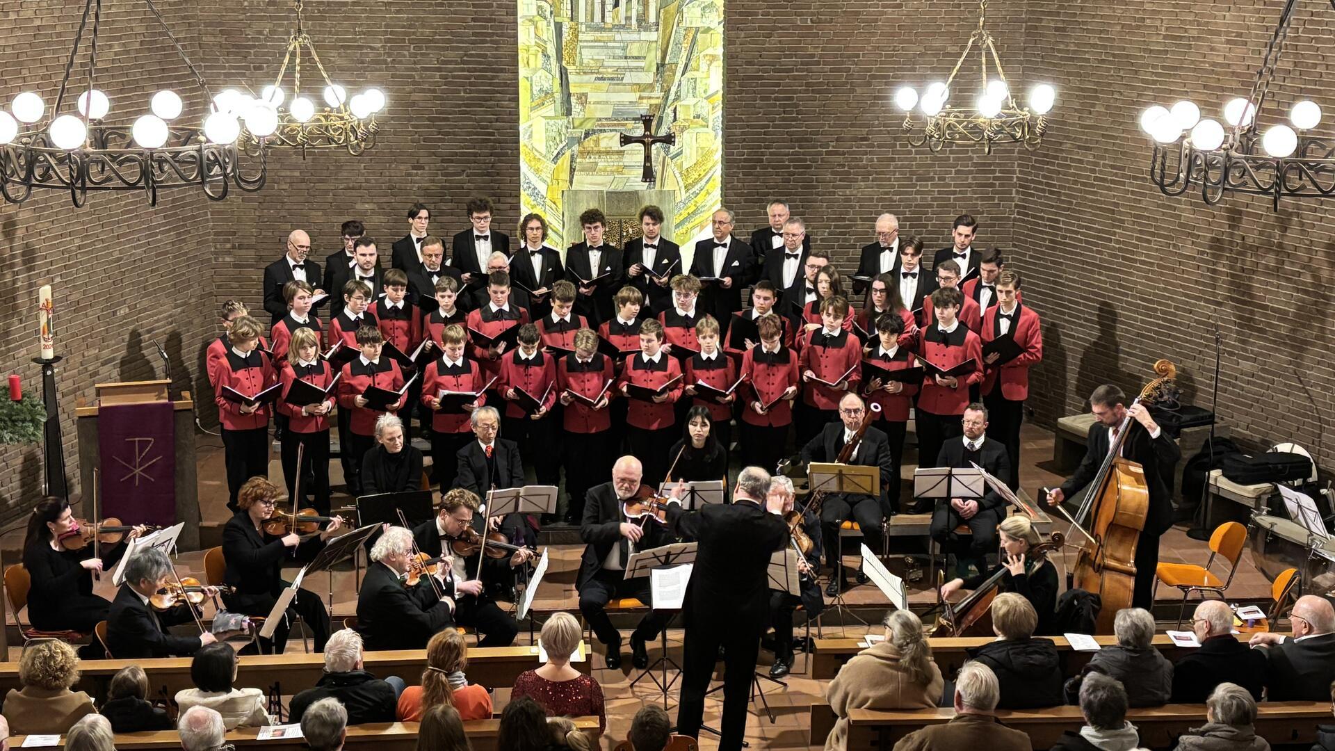 Ein Chor aus Männern und Jungen in schwarzer und roter Chorkleidung steht in einer Kirche vor einer Wand mit einem großen Mosaik. Vor dem Chor sitzt ein Orchester mit Musikern, die verschiedene Instrumente spielen, darunter Geigen, Cello und Kontrabass. Ein Dirigent steht in der Mitte und leitet das Ensemble. Im Vordergrund sind die Rücken von Zuschauern zu sehen, die das Konzert verfolgen.