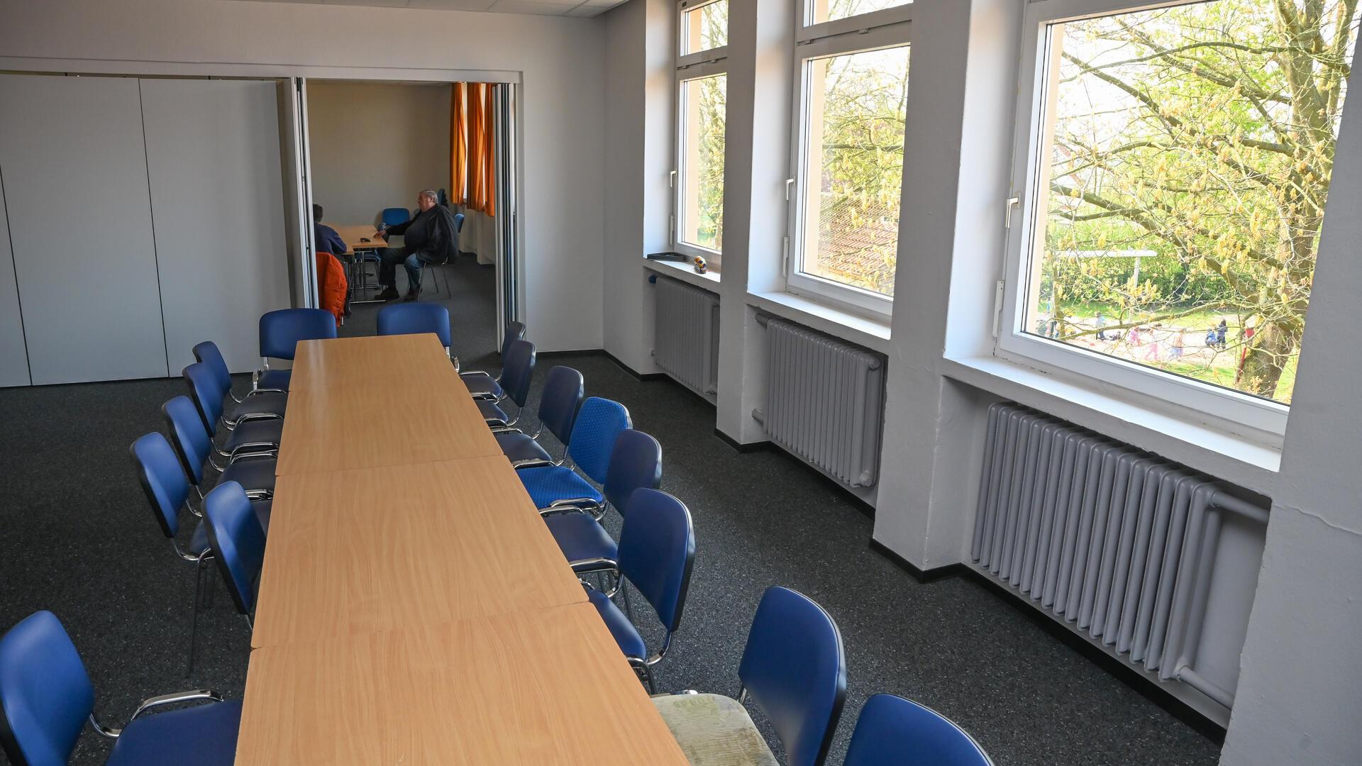 Ein renovierter Raum mit aneinander gestellten Tischen und blauen Stühlen. Ein Baum taucht hinter einem Fenster auf.