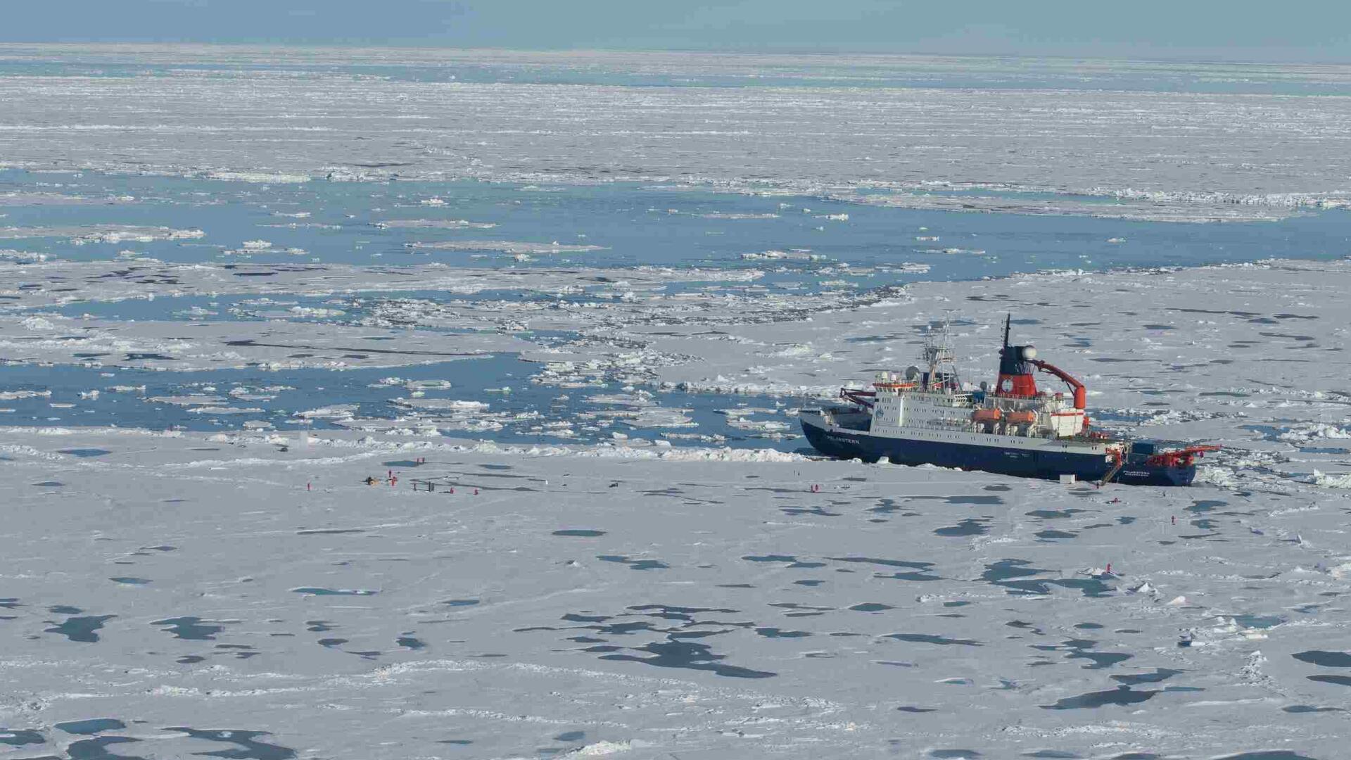 Der Forschungseisbrecher „Polarstern“ liegt inmitten von arktischem Meereis, umgeben von Schollen und offenen Wasserflächen. In der Nähe arbeiten Personen auf dem Eis an Messstationen.