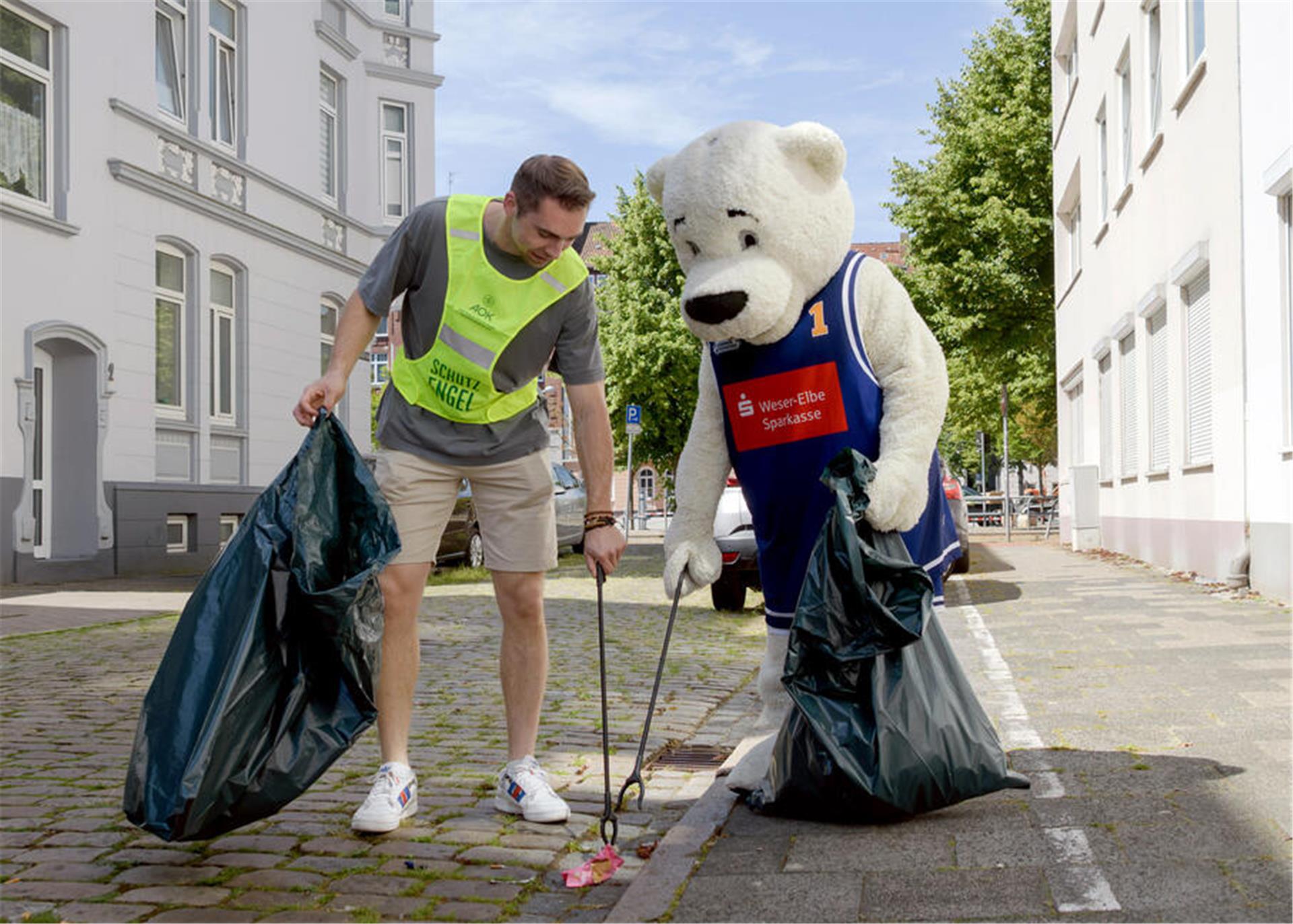 Ein Mann in gelbem Leibchen. Er sammelt mit einer Greifzange Müll auf. Ihm hilft ein "Eisbär", das Basketball-Maskottchen Dunky.