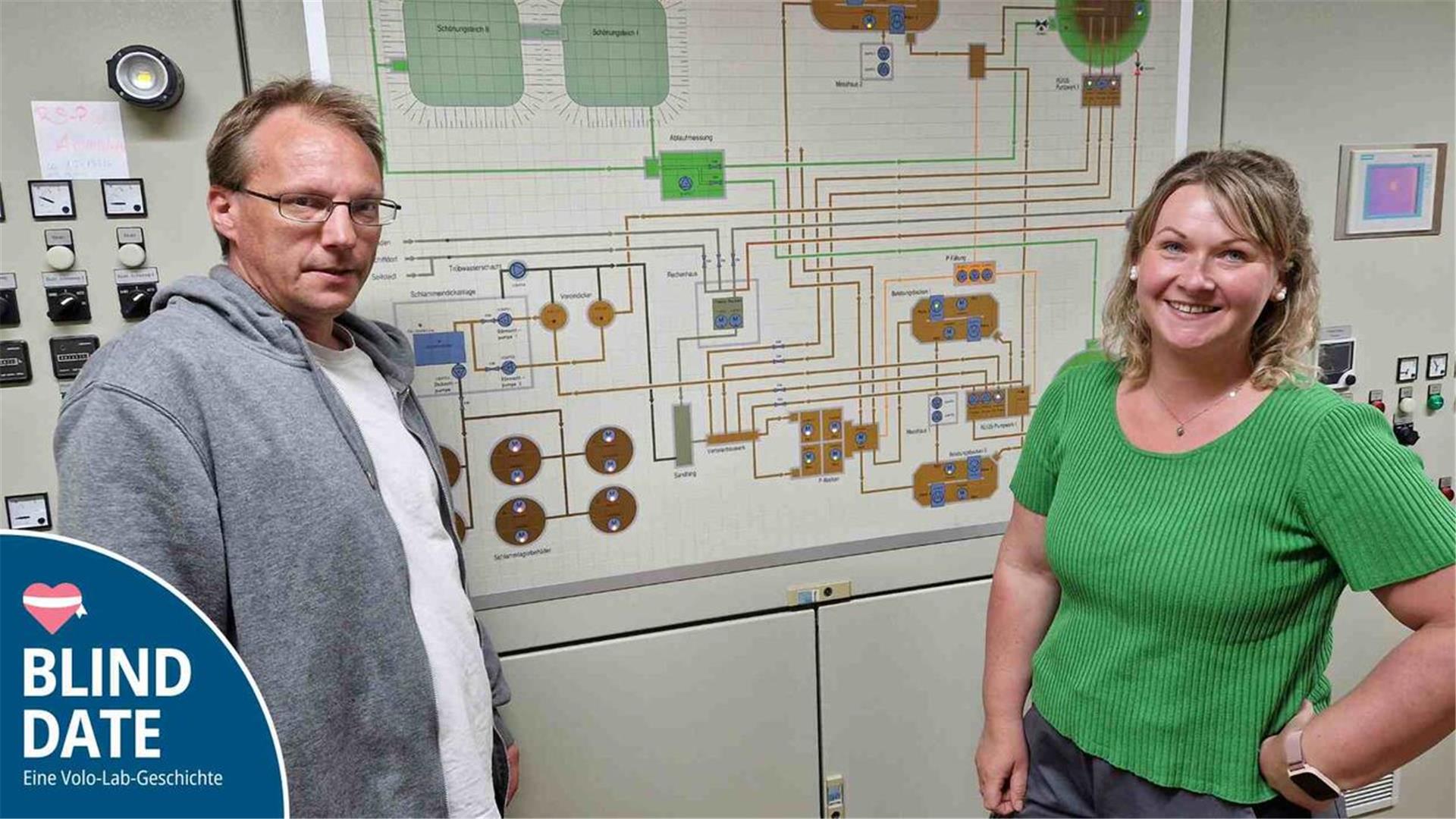 Ein Mann und eine Frau stehen vor einem großen Schaltplan an einer Wand.