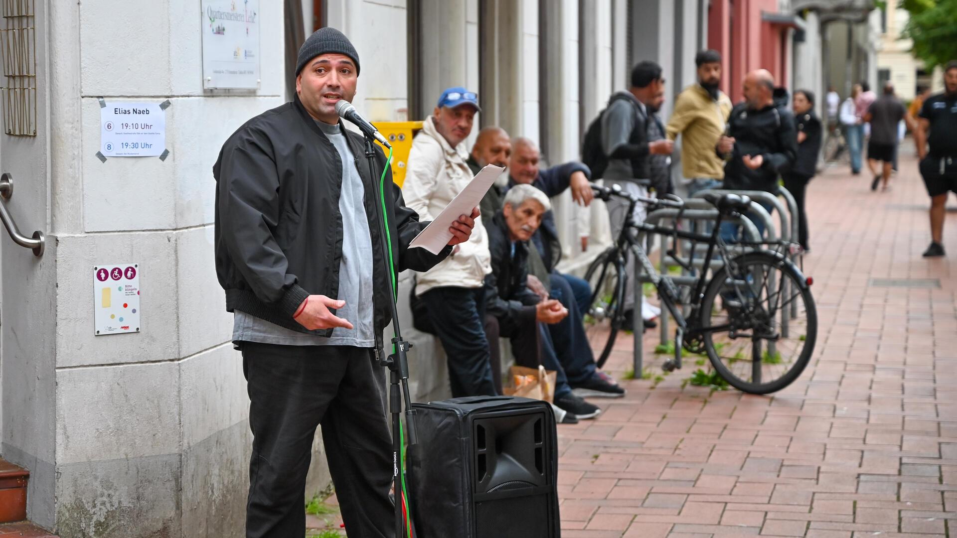 Poetry Slammer Elias Naeb trägt auf offener Straße vor, während Zuhörer auf einer Bank und Passanten aufmerksam lauschen.