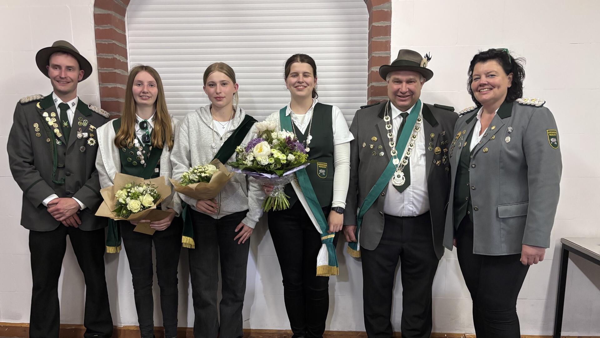Sechs Mitglieder eines Schützenvereins stehen nebeneinander vor einer weißen Wand mit Ziegelbögen. Sie tragen traditionelle Schützenuniformen mit Orden und Schärpen. Drei Frauen in der Mitte halten Blumensträuße. 