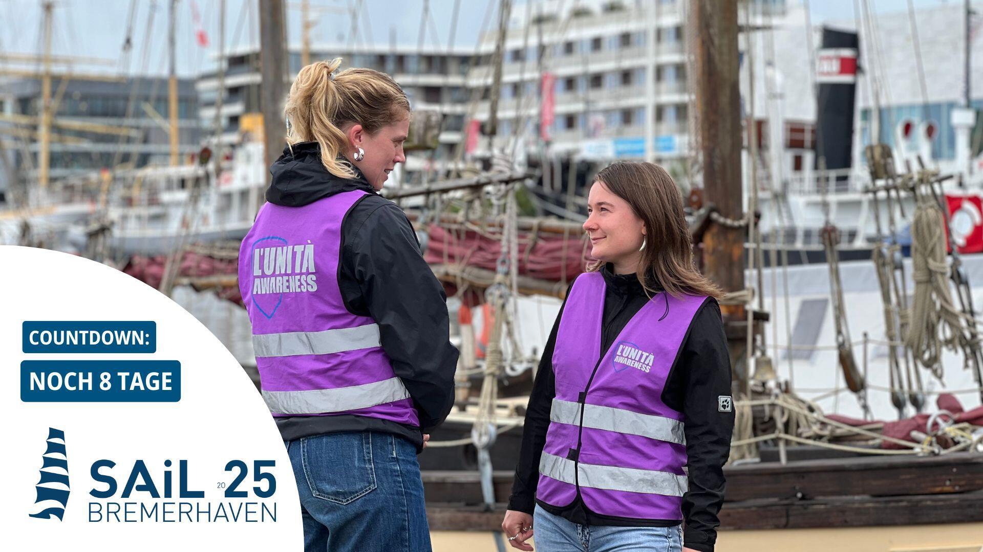 Zwei Frauen in violetten Westen stehen vor einem Schiff. Im Hintergrund Binnenhafen mit größeren Häusern.