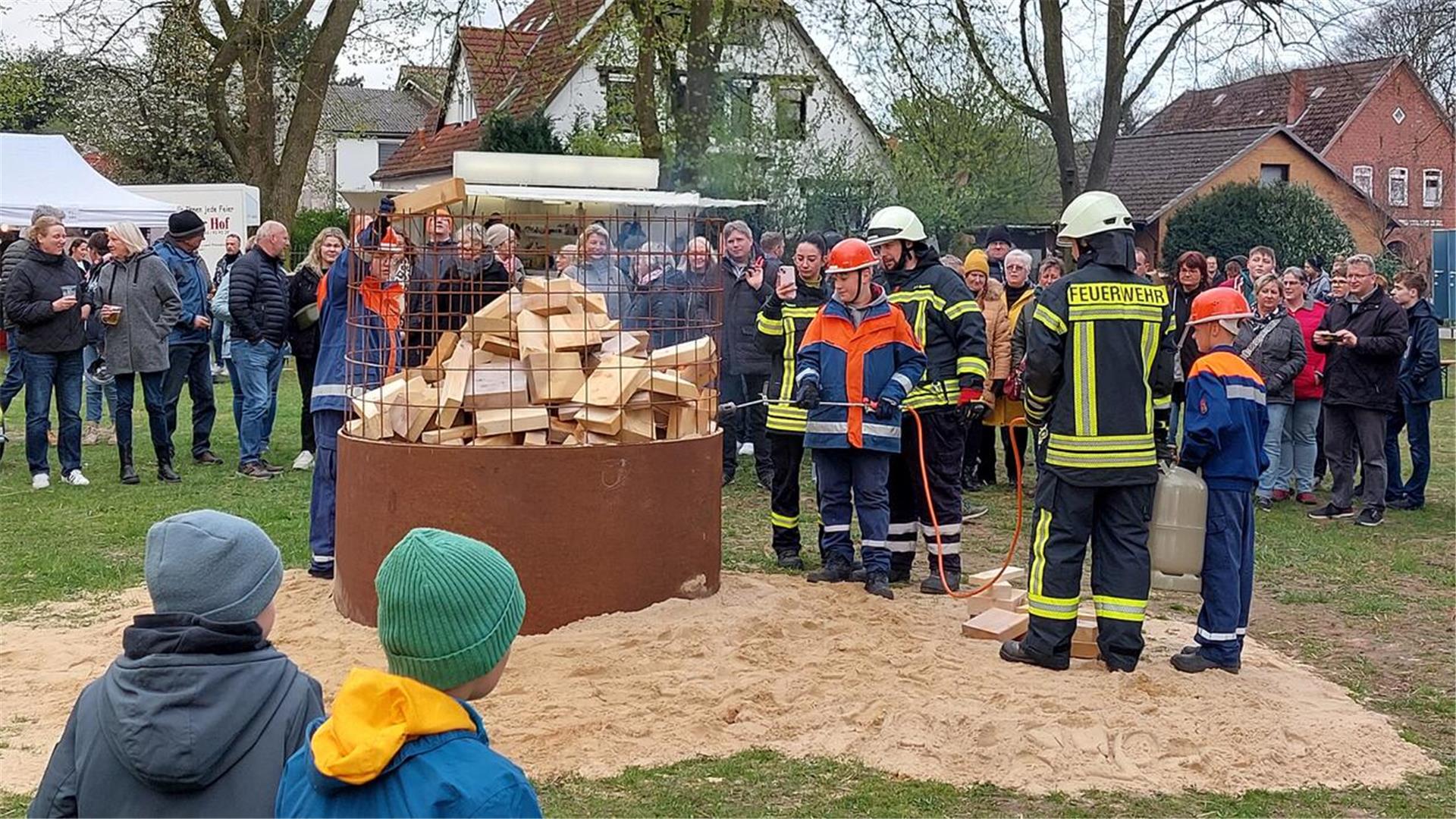 Jugendfeuerwehr und Feuerwehrleute entzünden unter Beobachtung zahlreicher Zuschauer ein Osterfeuer in einem Metallkorb mit Holzscheiten; Kinder schauen gespannt zu.