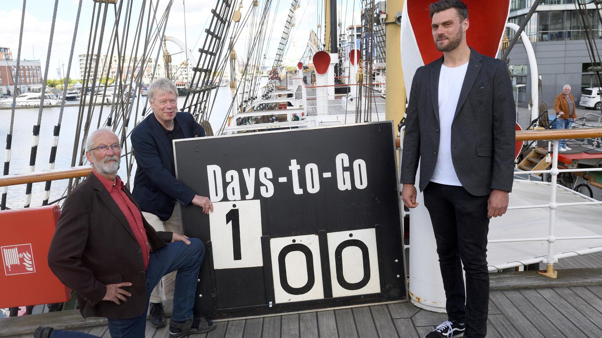 Drei Männer auf einem Schiff halten ein Schild in der Hand, auf dem „Days to go 100“ steht.