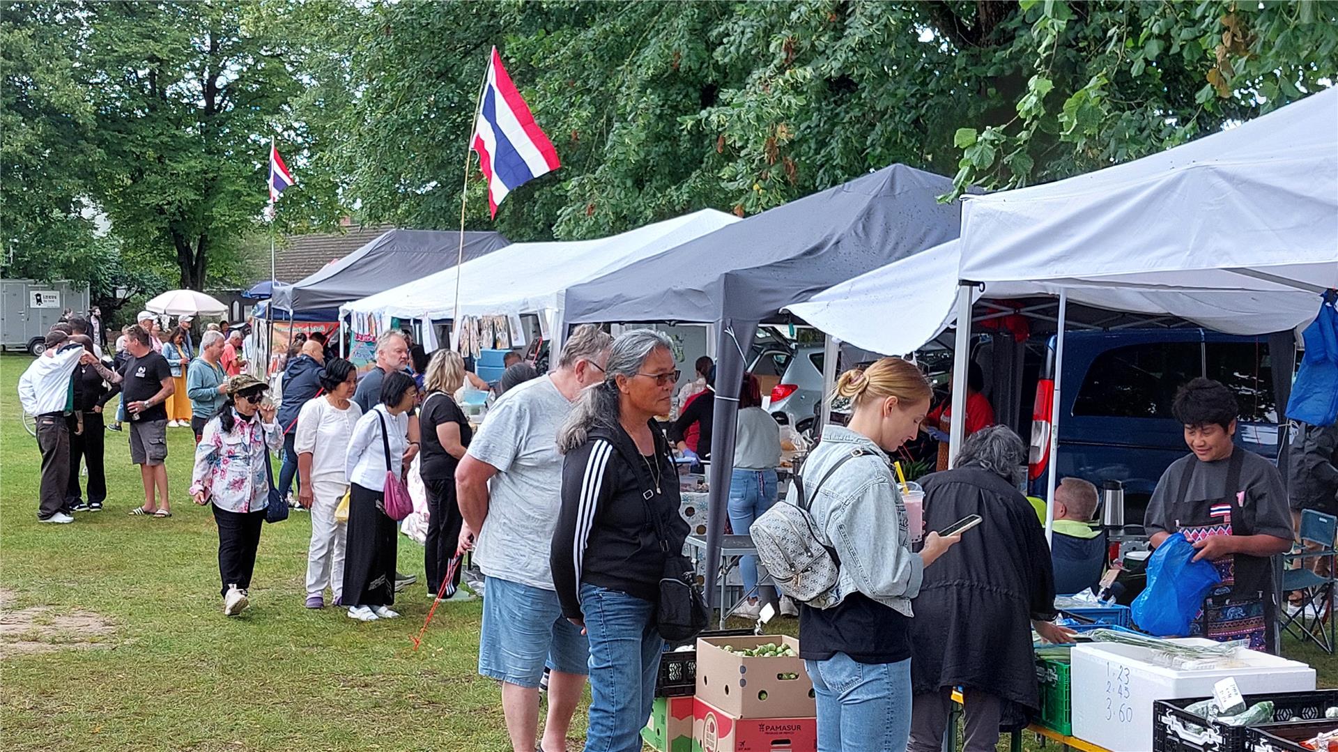 Besucher schlendern über das Thai-Kultur Streetfood Festival in Debstedt. Mehrere Verkaufsstände mit thailändischer Flagge bieten Speisen und Waren an, während Menschen in sommerlicher Kleidung die Stände begutachten.