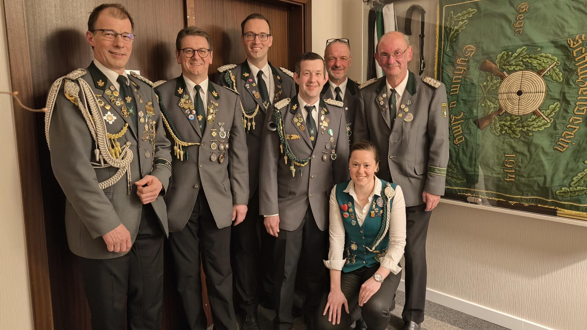 Sieben Mitglieder eines Schützenvereins posieren in festlicher Uniform vor einer Wand mit einer großen grünen Vereinsfahne. Die Männer tragen graue Jacken mit zahlreichen Orden und Abzeichen, eine Frau in der vorderen Reihe trägt eine grüne Weste mit Auszeichnungen.