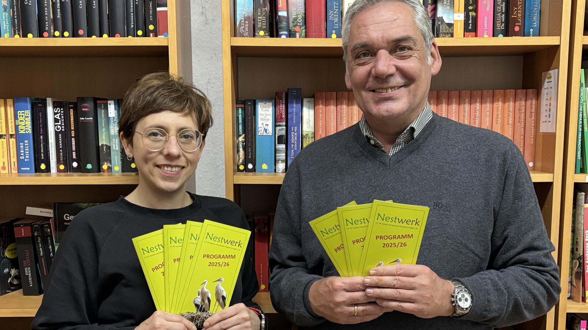 Eine Frau und ein Mann stehen vor einem Bücherregal und halten Flyer in den Händen.