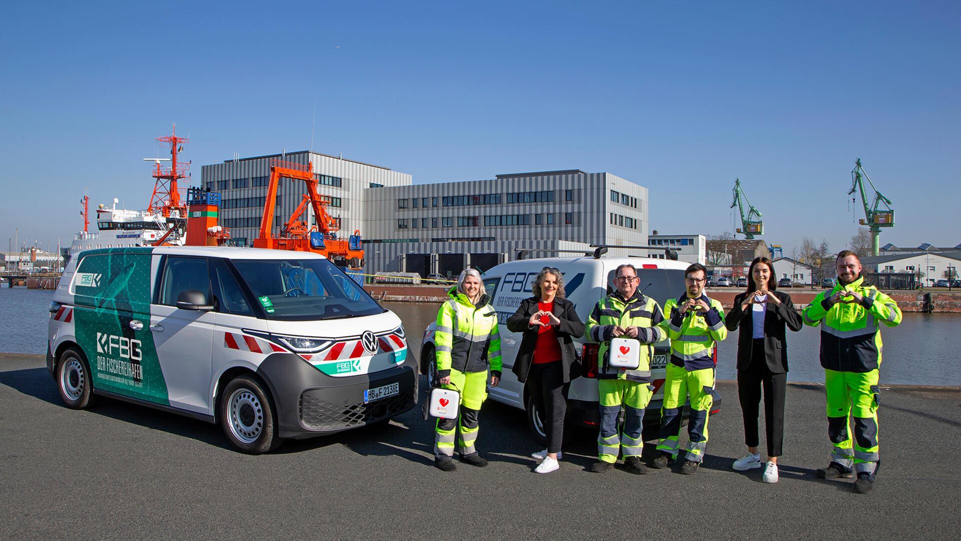 Eine Gruppe von Menschen, einige in auffälliger Arbeitskleidung, steht vor zwei Transportern mit der Aufschrift „FBG“. Die Transporter sind auf einem befestigten Bereich in der Nähe eines Hafens mit Kränen und Industriegebäuden im Hintergrund geparkt. Die Menschen formen Herzzeichen mit ihren Händen, und eine Person hält einen Erste-Hilfe-Kasten mit einem Herzsymbol darauf. Die Szene deutet auf einen Fokus auf Sicherheits- oder Notfallmaßnahmen hin.