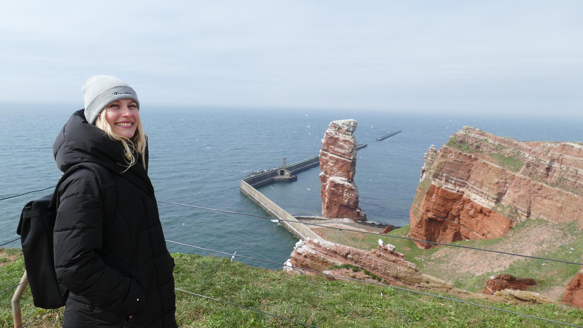 Eine junge Frau mit blonden Haaren steht auf dem Helgoländer Oberland vor der Küste. Sie trägt eine dicke schwarze Jacke und eine graue Mütze. Im Hintergrund sind der Helgoländer Felsen "Lange Anna" und die Nordsee zu sehen.