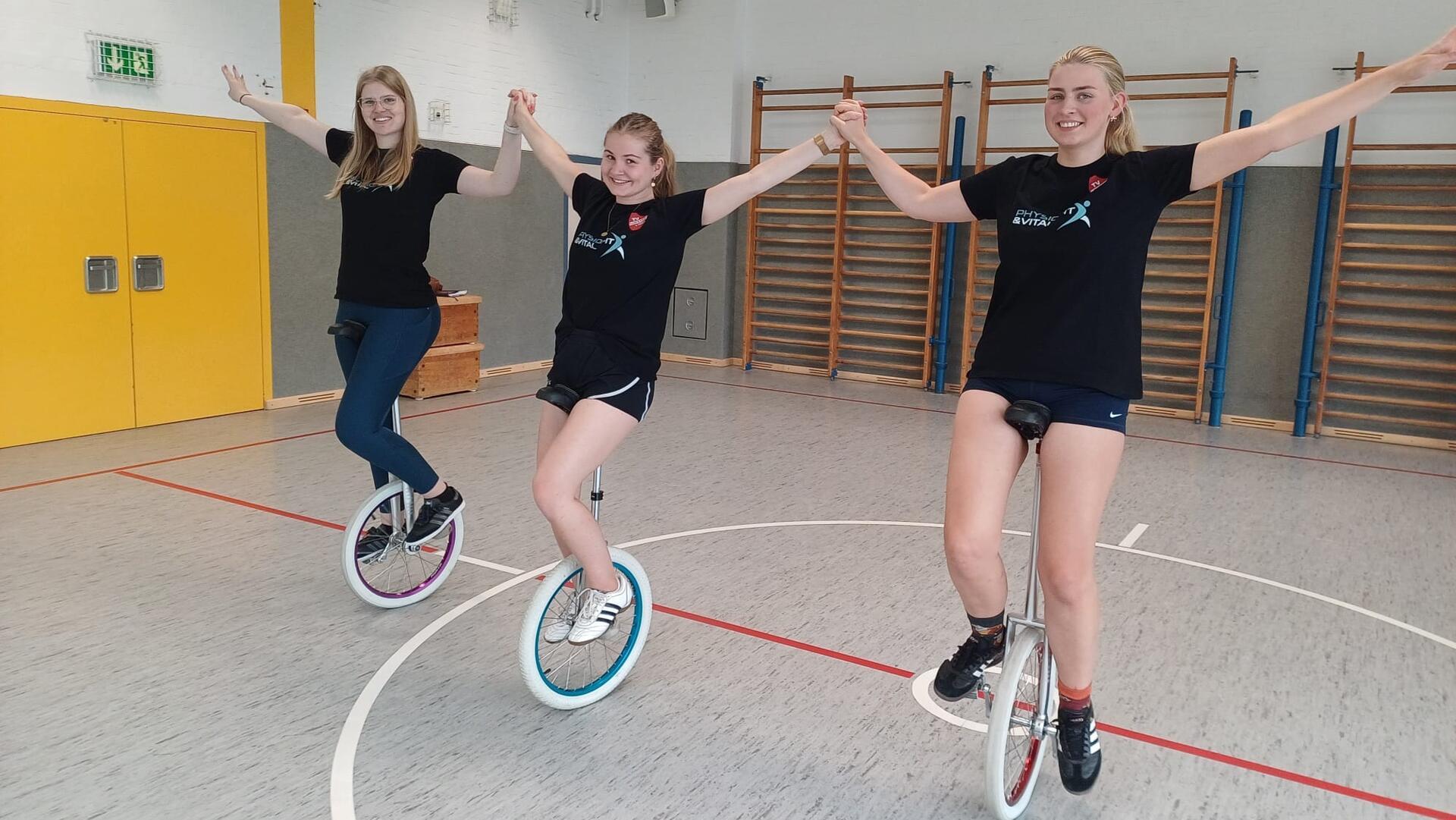 Drei junge Frauen auf Einrädern in einer Halle. 