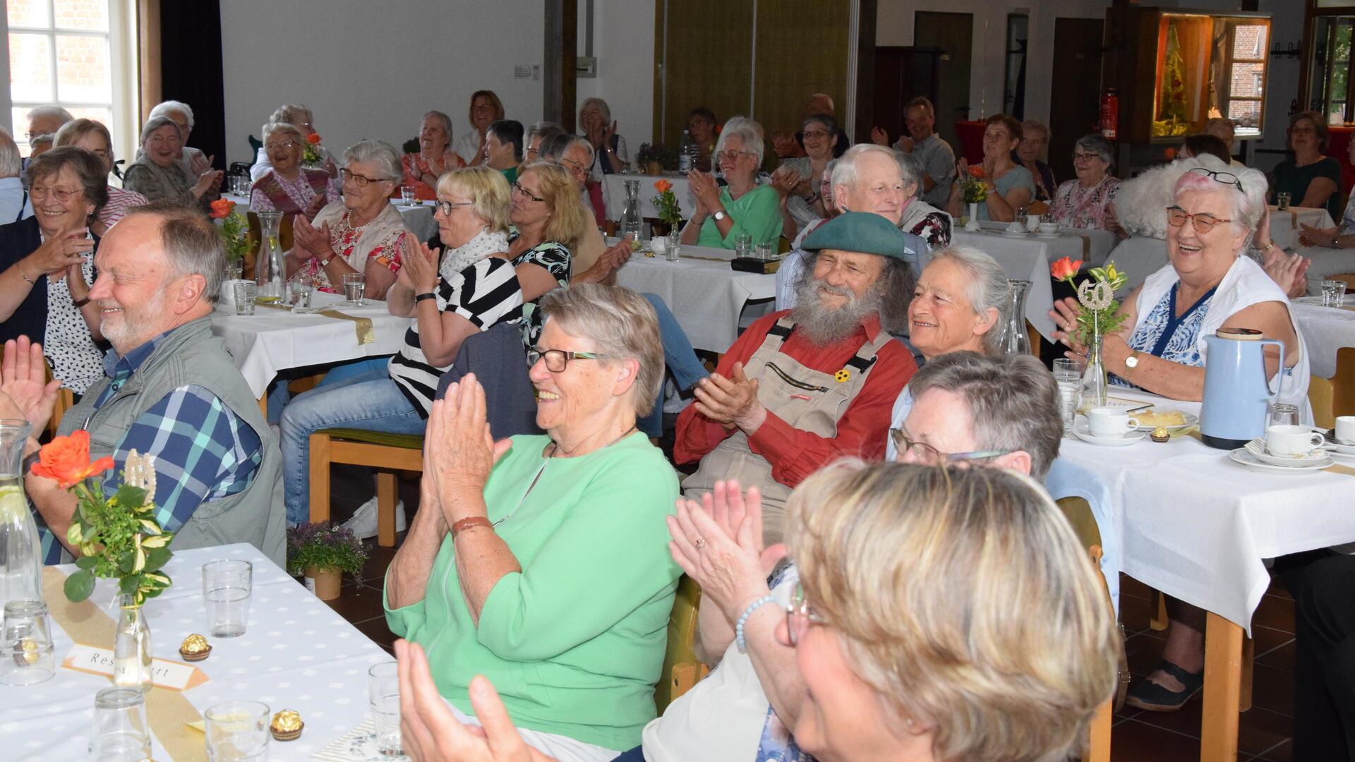 Ältere Frauen und Männer sitzen an Tischen, lachen und applaudieren.