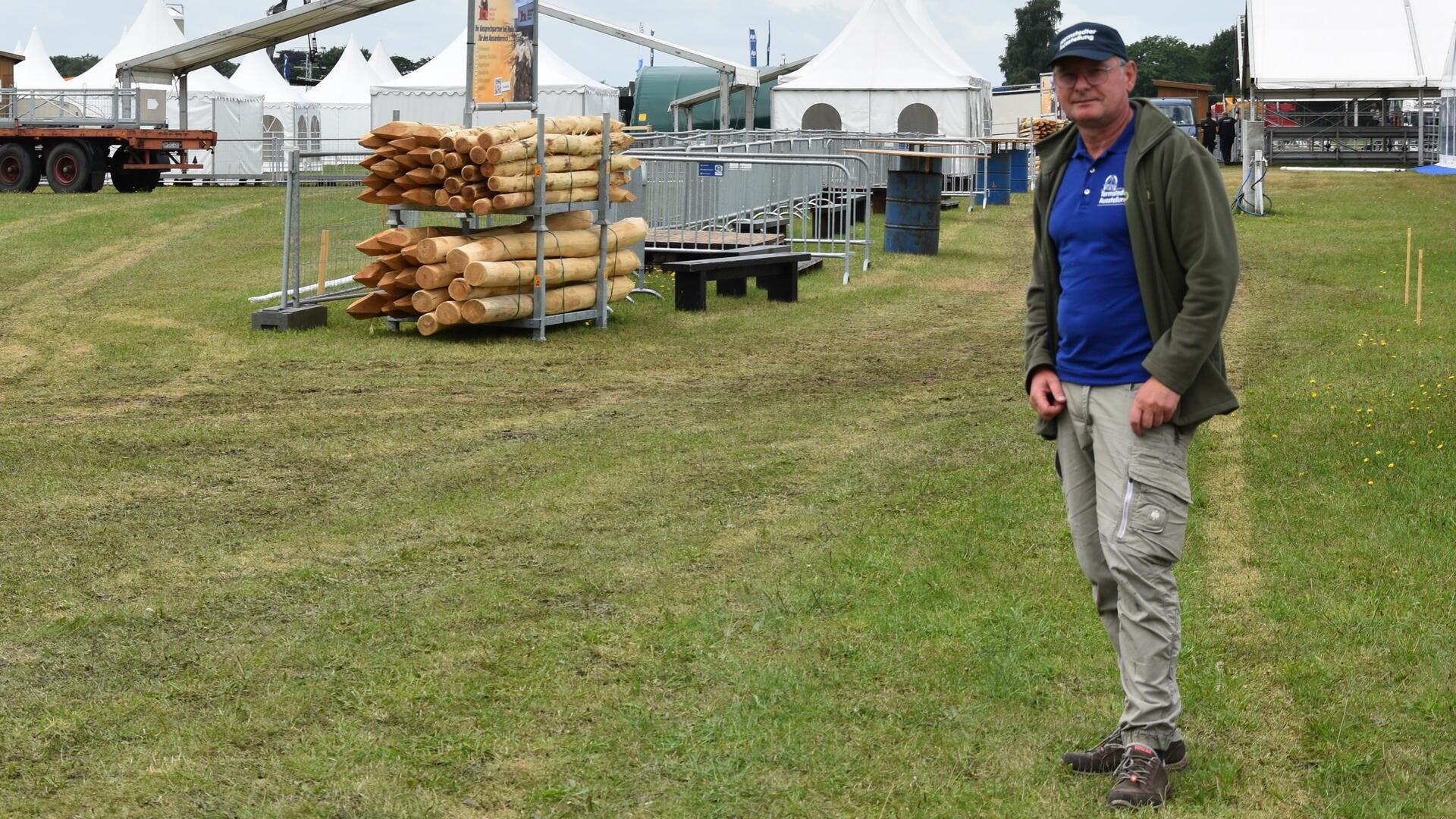 Ein älterer Mann in blauem T-Shirt steht auf einer Wiese. Um ihn herum werden Zelte und Zäune errichtet.