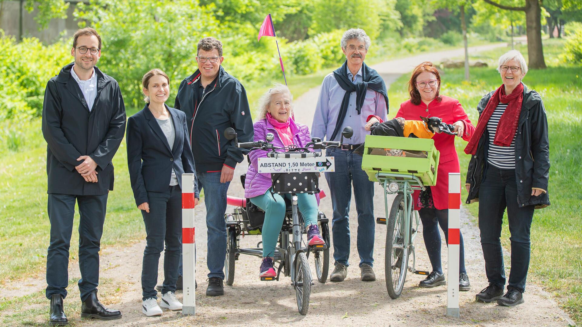 Eine Gruppe von sieben Erwachsenen steht auf einem Rad- und Spazierweg im Grünen. Die Frau in der Mitte sitzt auf einem Dreiradtandem mit Schild "Abstand 1,50 Meter". Rechts neben ihr steht eine Frau mit rotem Mantel und Fahrrad mit grünem Transportkorb, in dem ein kleiner Hund sitzt.
