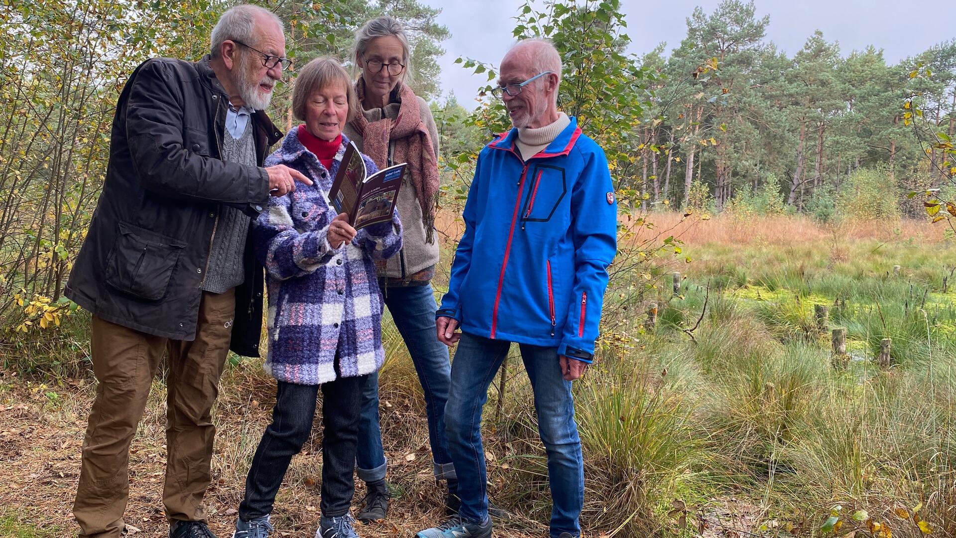 Vier Menschen stehen in einer Moorlandschaft und betrachten ein Buch. 
