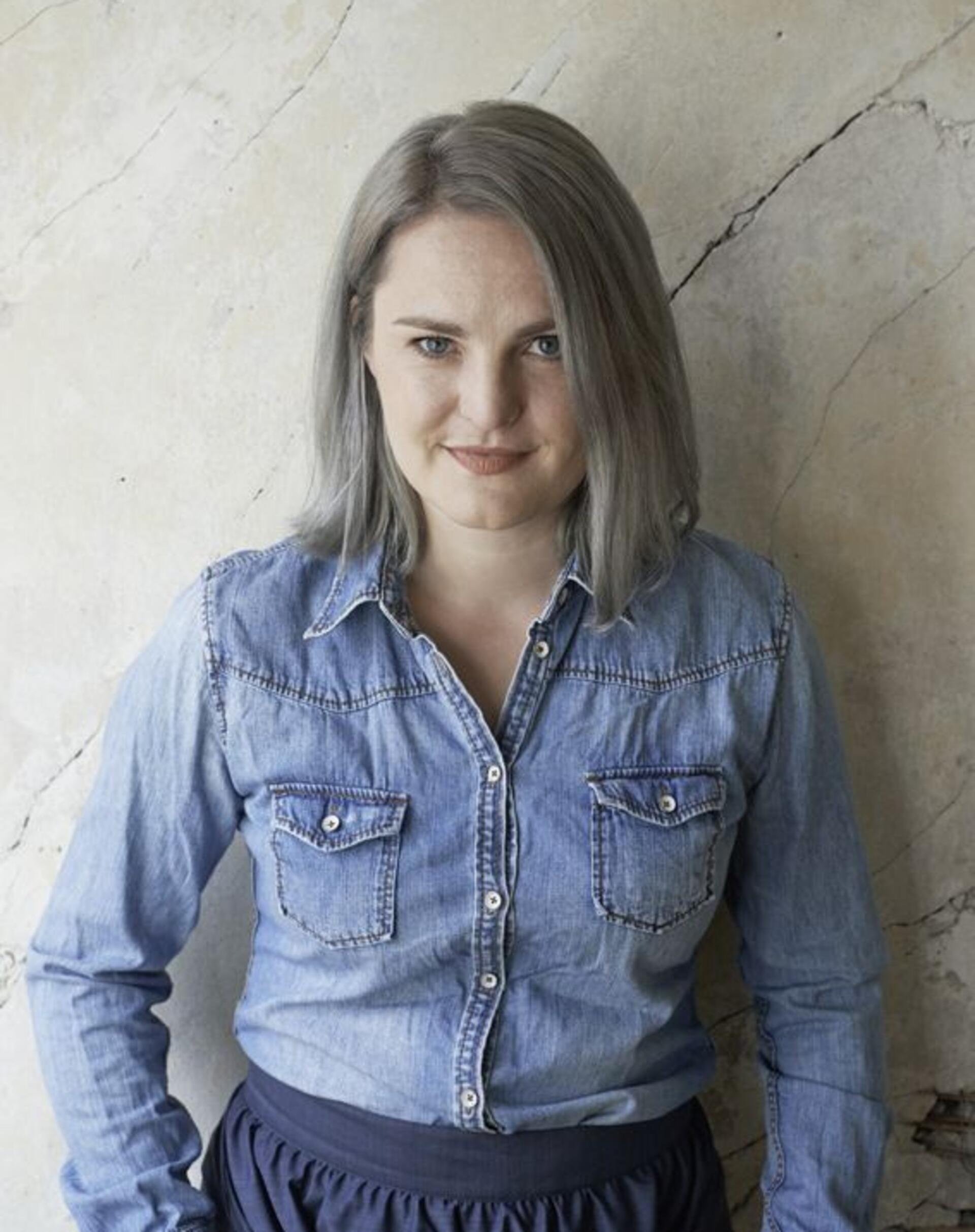 Masche für Masche: Friederike „Fredda“ Hansmann liest in Zeven Eine Frau mit halblangen Haaren in einem Jeanshemd steht vor einer Wand.