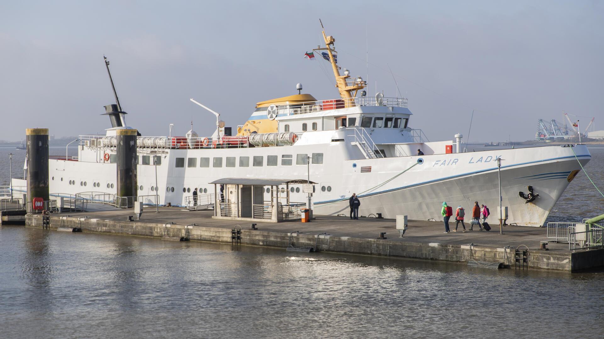 Ein Schiff liegt an der Kaje. Ein paar Fahrgäste sind mit Rucksäcken und Koffer im Anmarsch. 