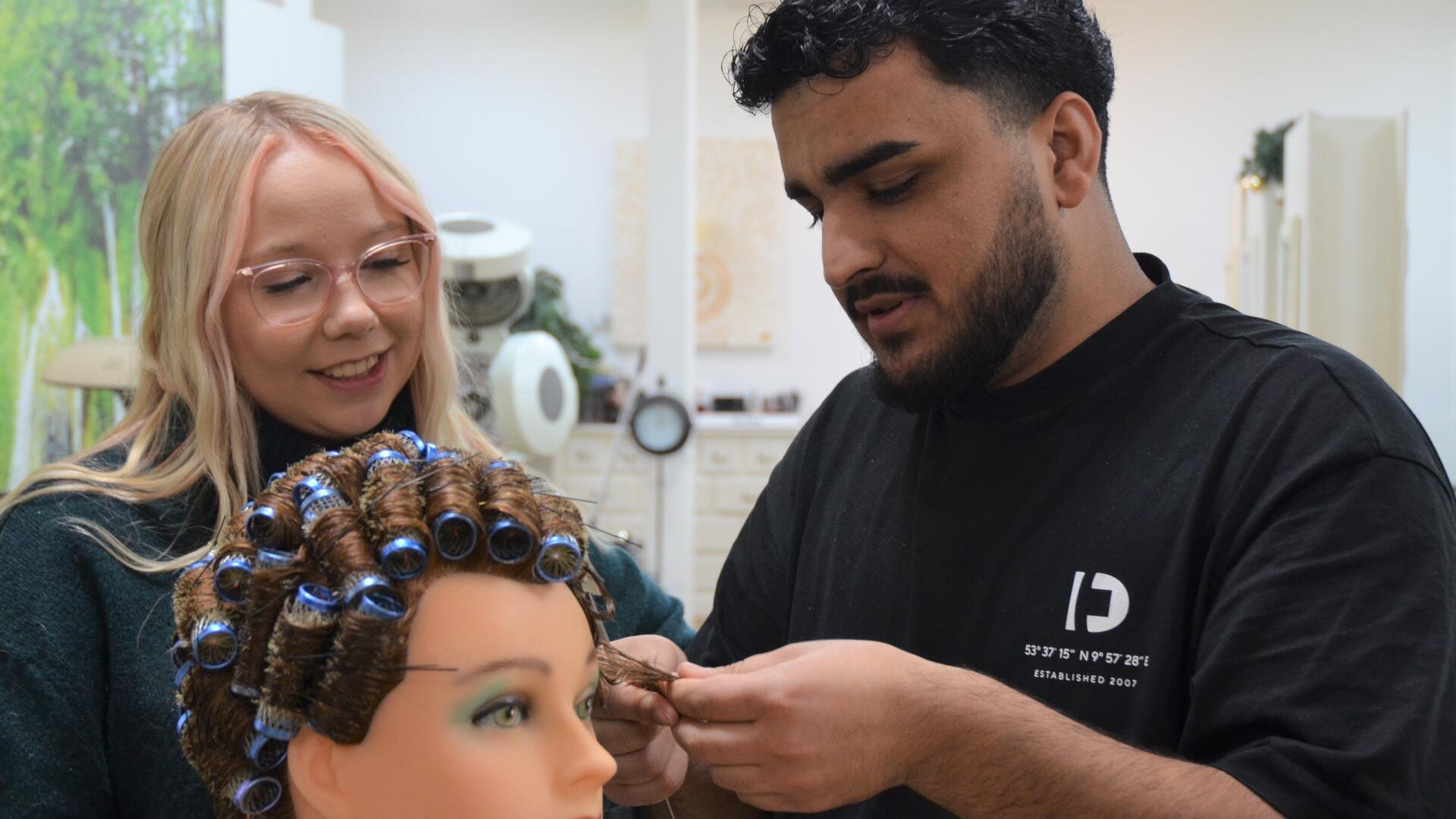 Ein Mann dreht die Haare einer Friseurpuppe mit Lockenwicklern auf. Eine Frau schaut ihm dabei zu. 