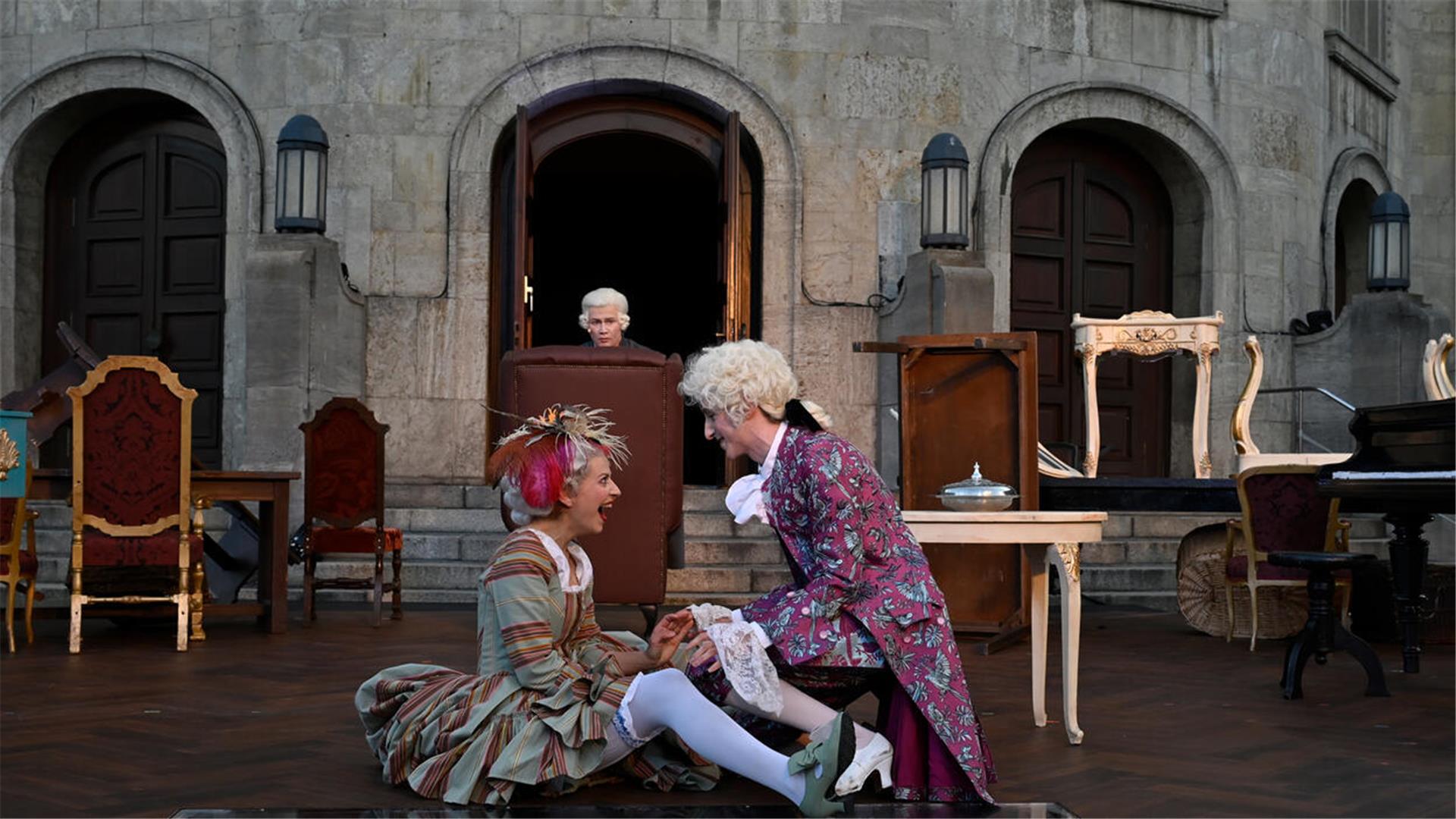Auf einer Theaterbühne drei Personen in barocken Kostümen. Eine Frau und ein Mann im Vordergrund albern herum, im Hintergrund ein Mann im Ohrensessel.