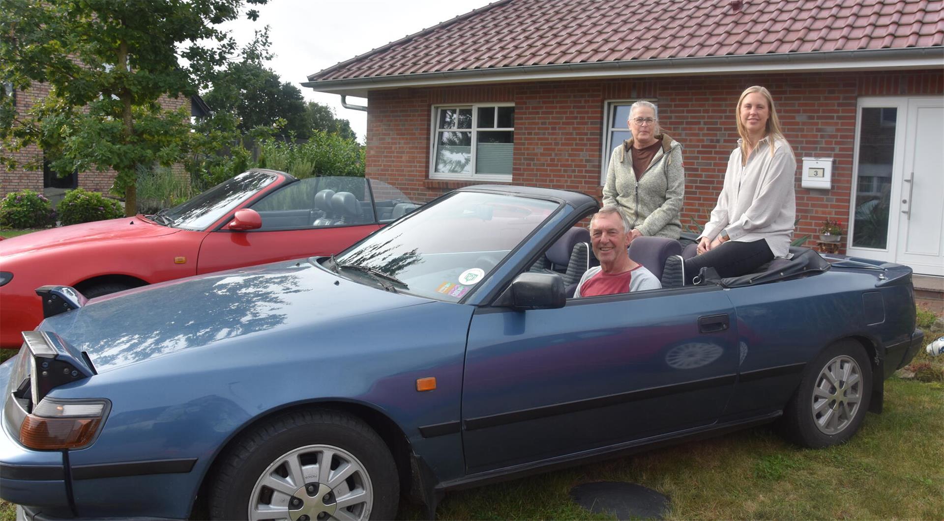 Ein Mann sitzt am Steuer eines Cabriolets, hinten auf dem Verdeck sitzen zwei Frauen.