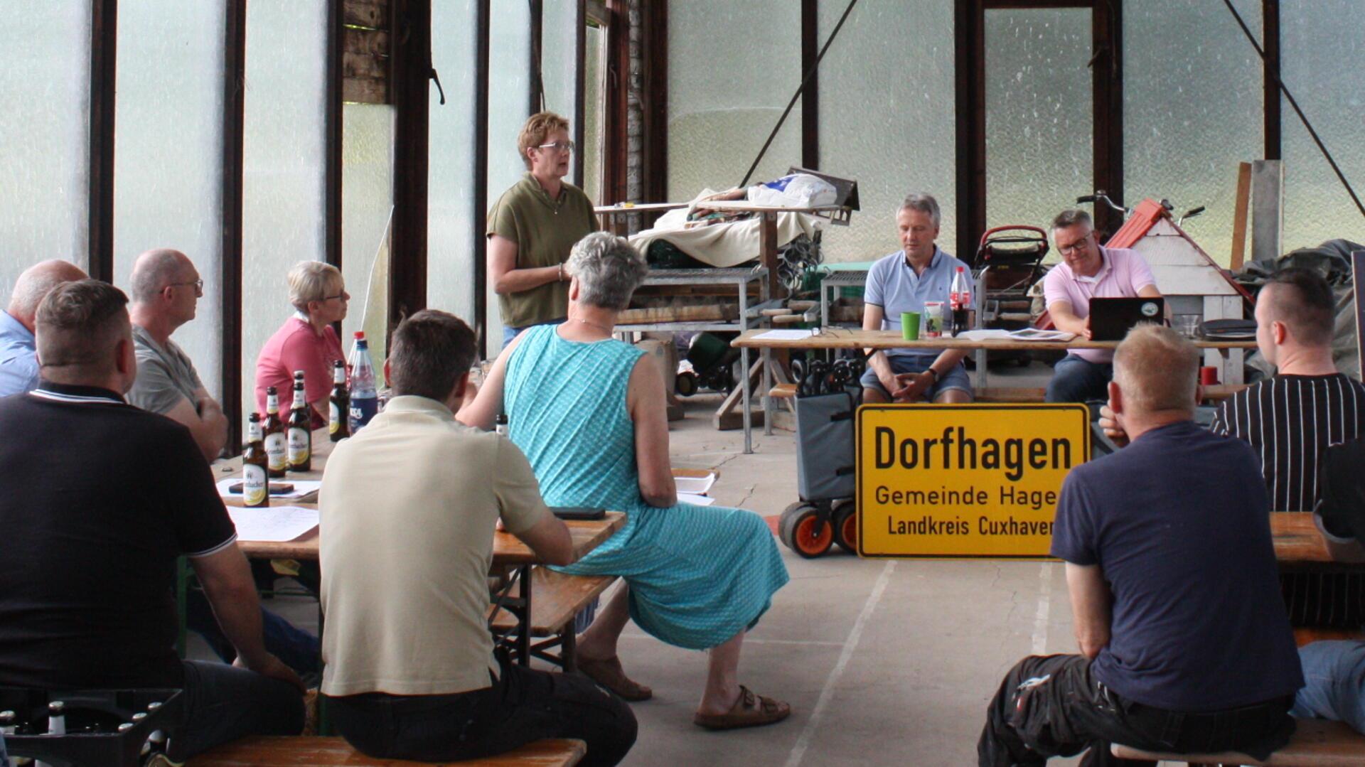 Einwohnerversammlung in Dorfhagen: Mehrere Personen sitzen an Tischen in einer Halle, eine Frau steht und spricht, im Vordergrund ein Ortsschild von Dorfhagen.
