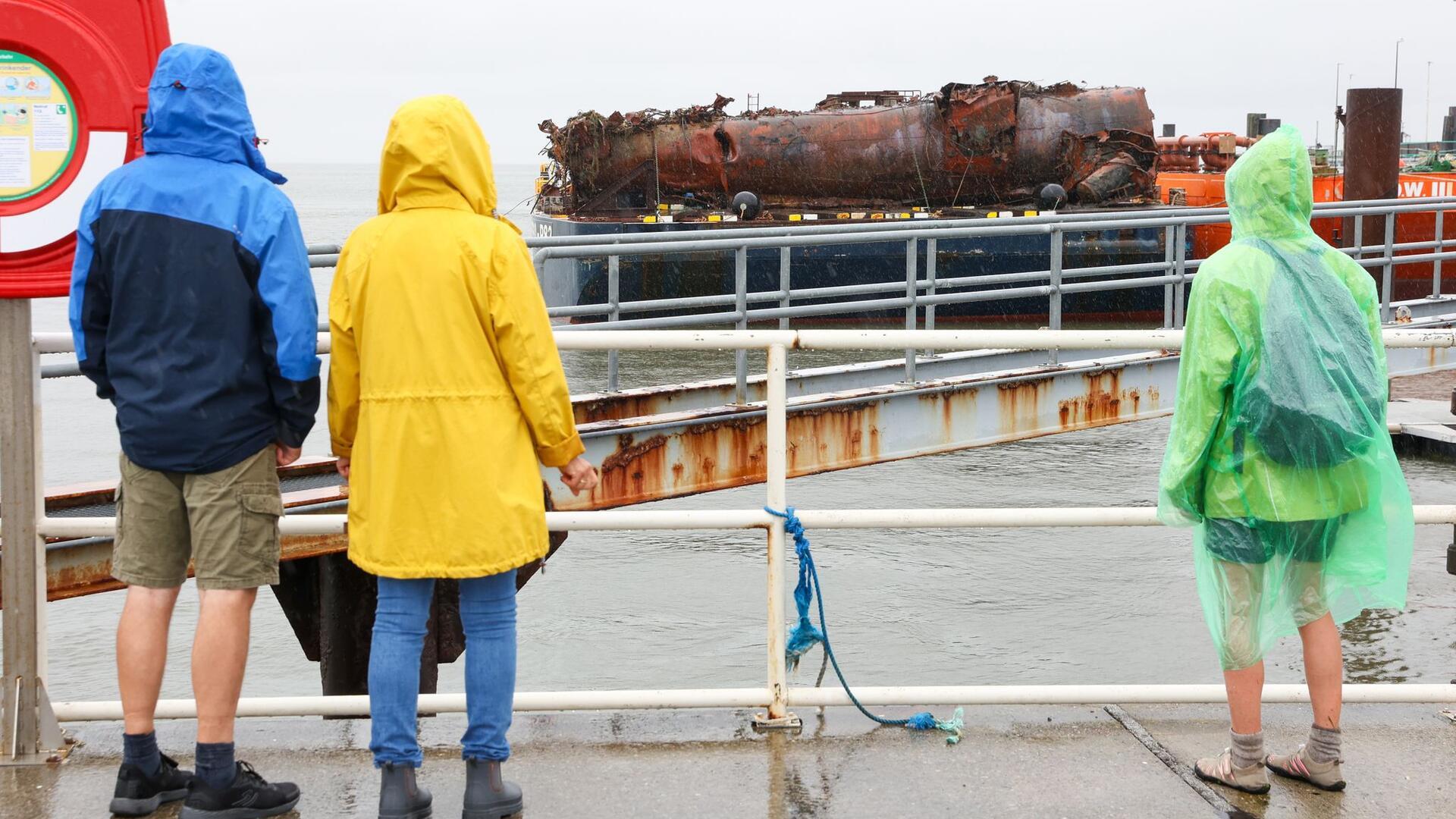 Drei Personen in Regenkleidung beobachten ein Schiffswrack, das auf einem Schiff angeliefert wird