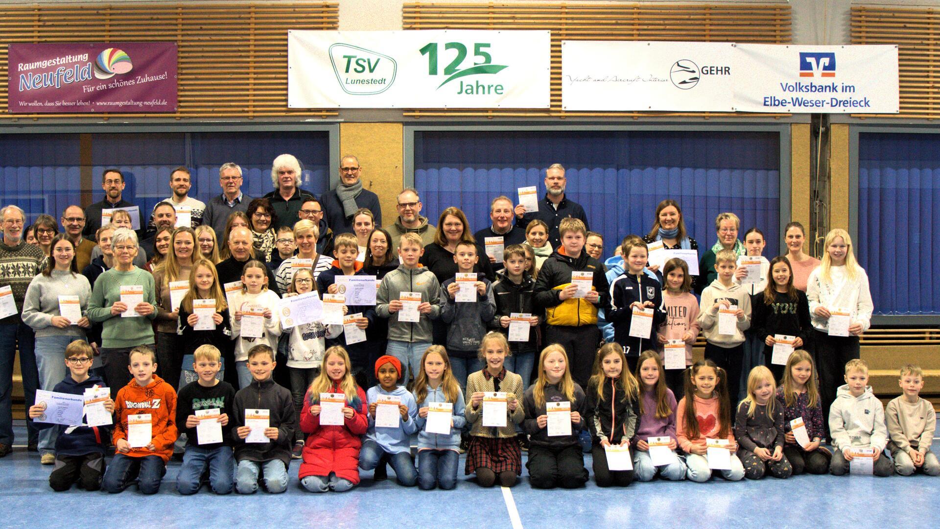 Kinder und Erwachsene knien und stehen in einer Sporthalle. In ihren Händen halten sie Urkunden.