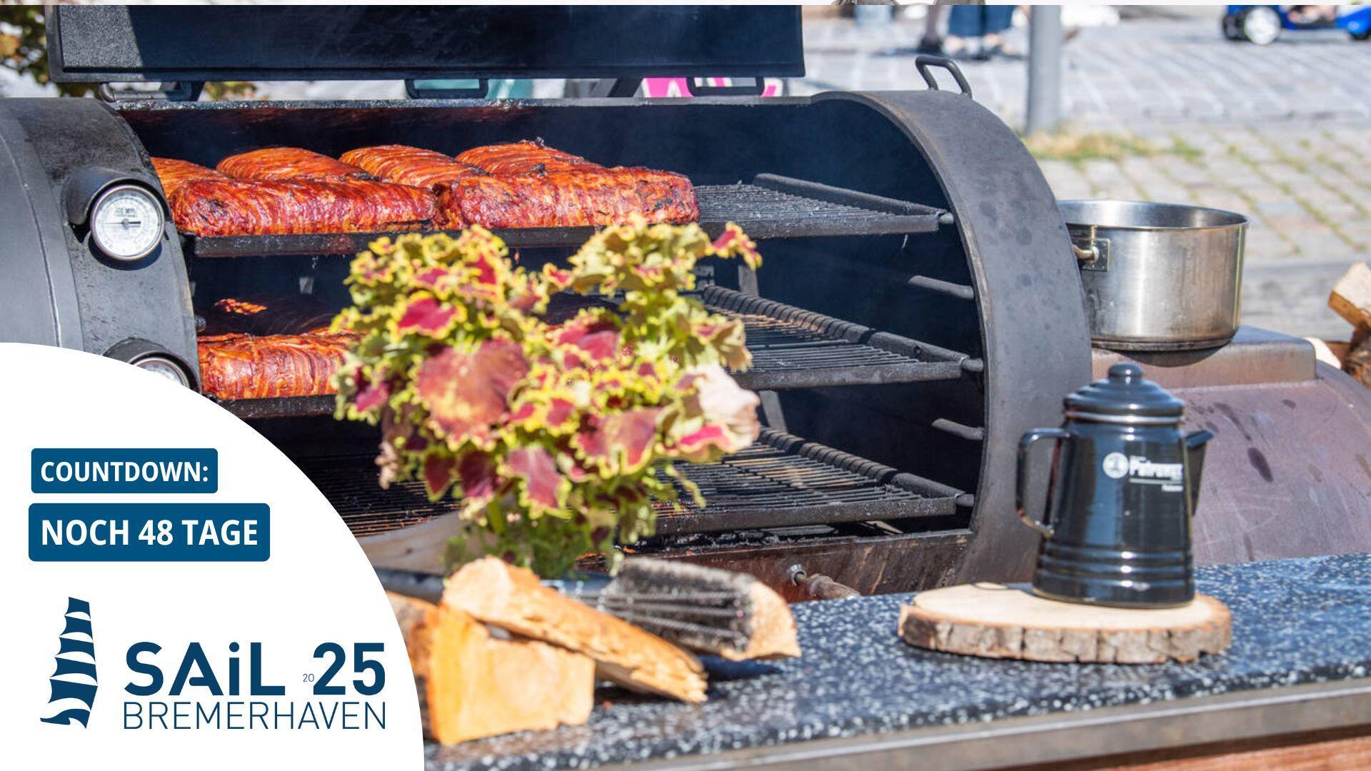Ein Grill, auf dem Fleisch liegt. Iim Vordergrund eine Pflanze, eine Kanne und Holzstücke auf einem Tresen.