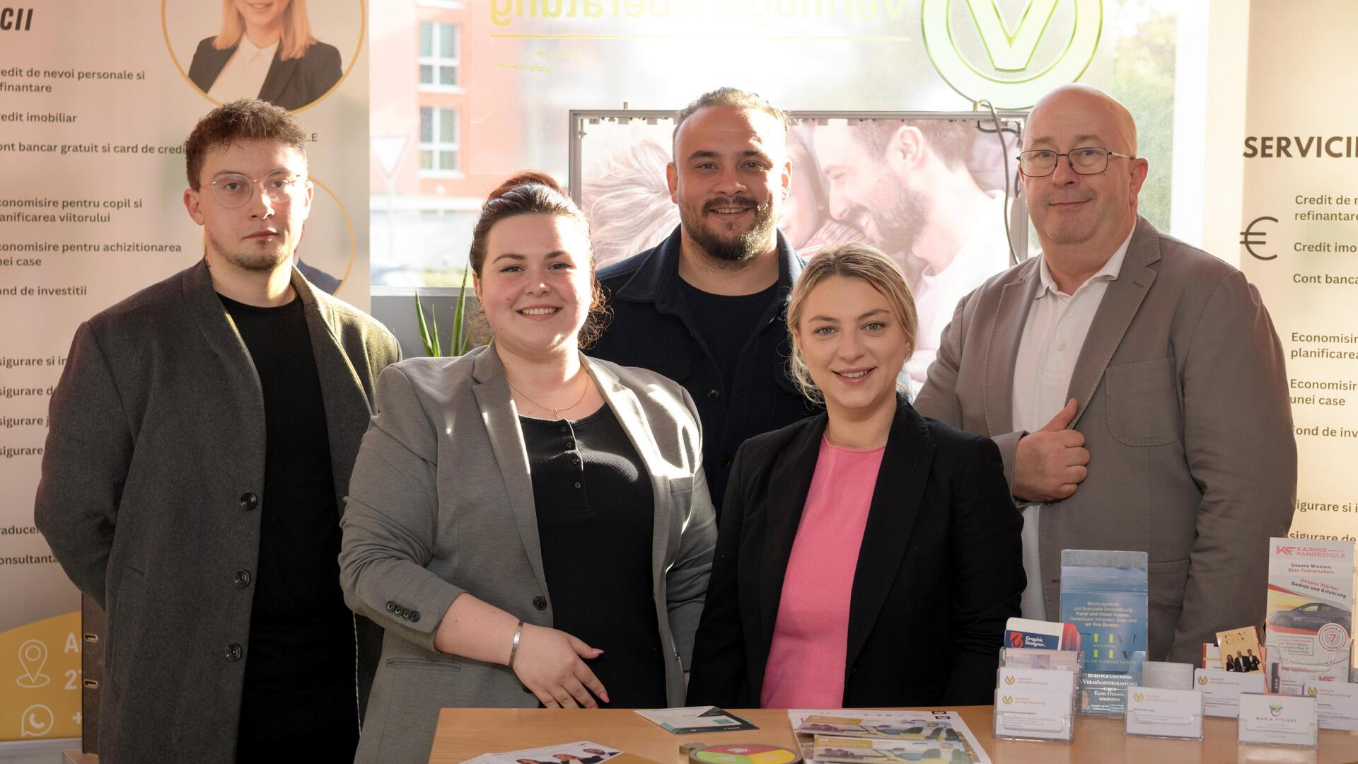 Fünf Personen – ein Mann, eine Frau, ein Mann, eine Frau und ein Mann - stehen an einem Tisch. Sie gucken in die Kamera.