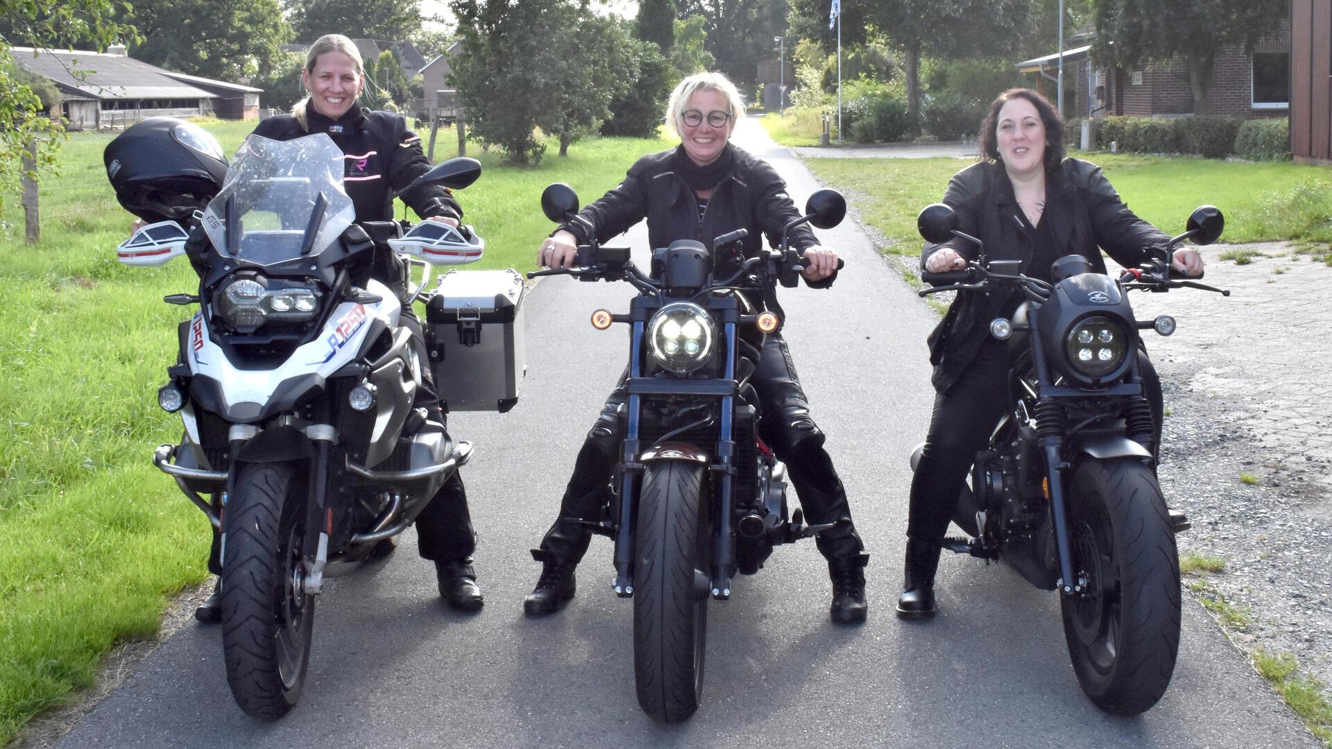 Drei Frauen auf Motorrädern stehen nebeneinander auf einer Straße.