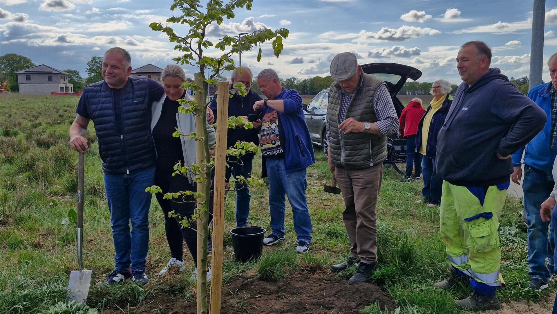 Mehrere Personen pflanzen gemeinsam einen jungen Baum auf einer Wiese in Lunestedt; im Hintergrund Neubauten, Autos und weitere Helfer und Zuschauer bei der Pflanzaktion.