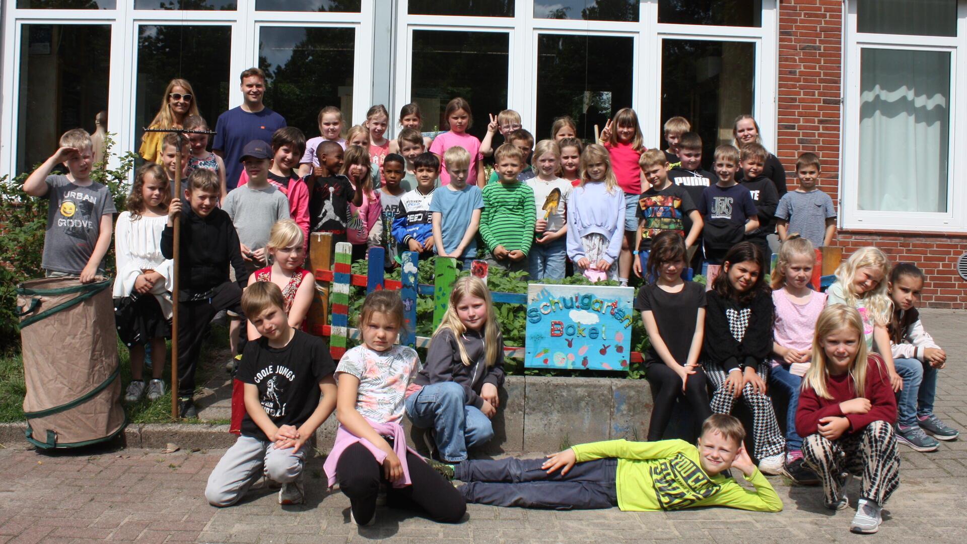 Gruppenfoto von etwa 45 Grundschulkindern und zwei Erwachsenen vor dem Schulgarten der Grundschule Bokel. Die Kinder stehen und sitzen um bunte Beete herum, im Vordergrund ist ein bemaltes Schild mit der Aufschrift „Schulgarten Bokel“ zu sehen.