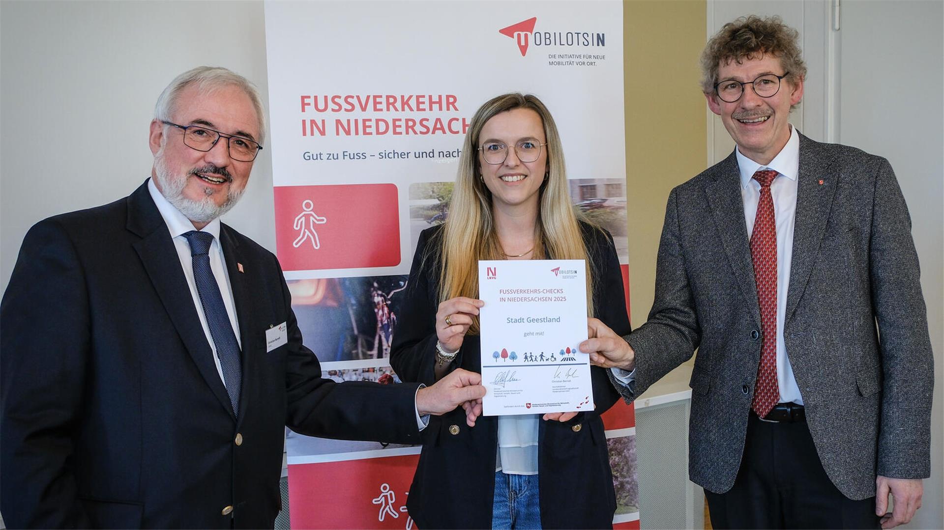 **ALT-Text:**  
Drei Personen bei der Urkundenübergabe zum Fußverkehrs-Check Niedersachsen; eine Frau in der Mitte hält die Urkunde der Stadt Geestland, flankiert von zwei Männern im Anzug, im Hintergrund ein Roll-up mit der Aufschrift „Fußverkehr in Niedersachsen“.