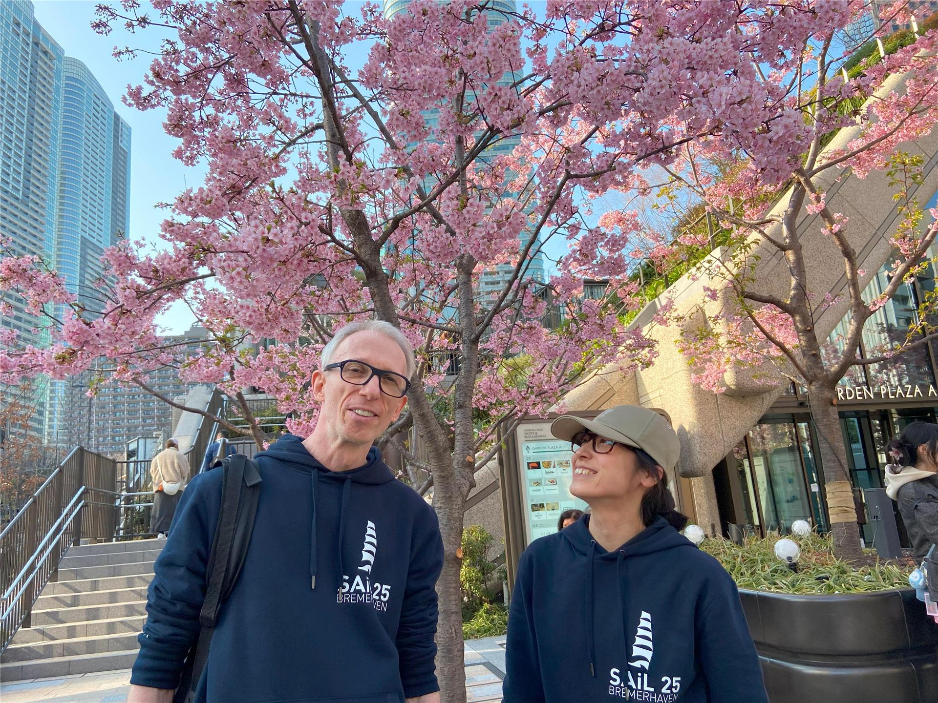 Ein Mann (links) und eine Frau stehen in Tokia vor einem Baum und tragen einen Hoodie mit der Aufschrift Sail. 