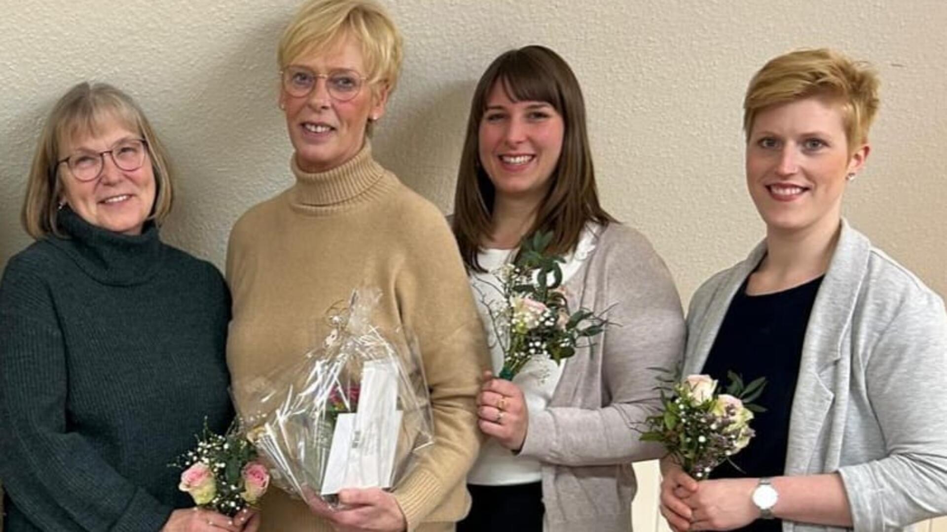 Vier Frauen stehen nebeneinander vor einer beigen Wand und lächeln in die Kamera. Die Frauen halten kleine Blumensträuße mit rosa und weißen Blüten, während die zweite Frau von links zusätzlich ein Geschenk in transparenter Verpackung trägt.