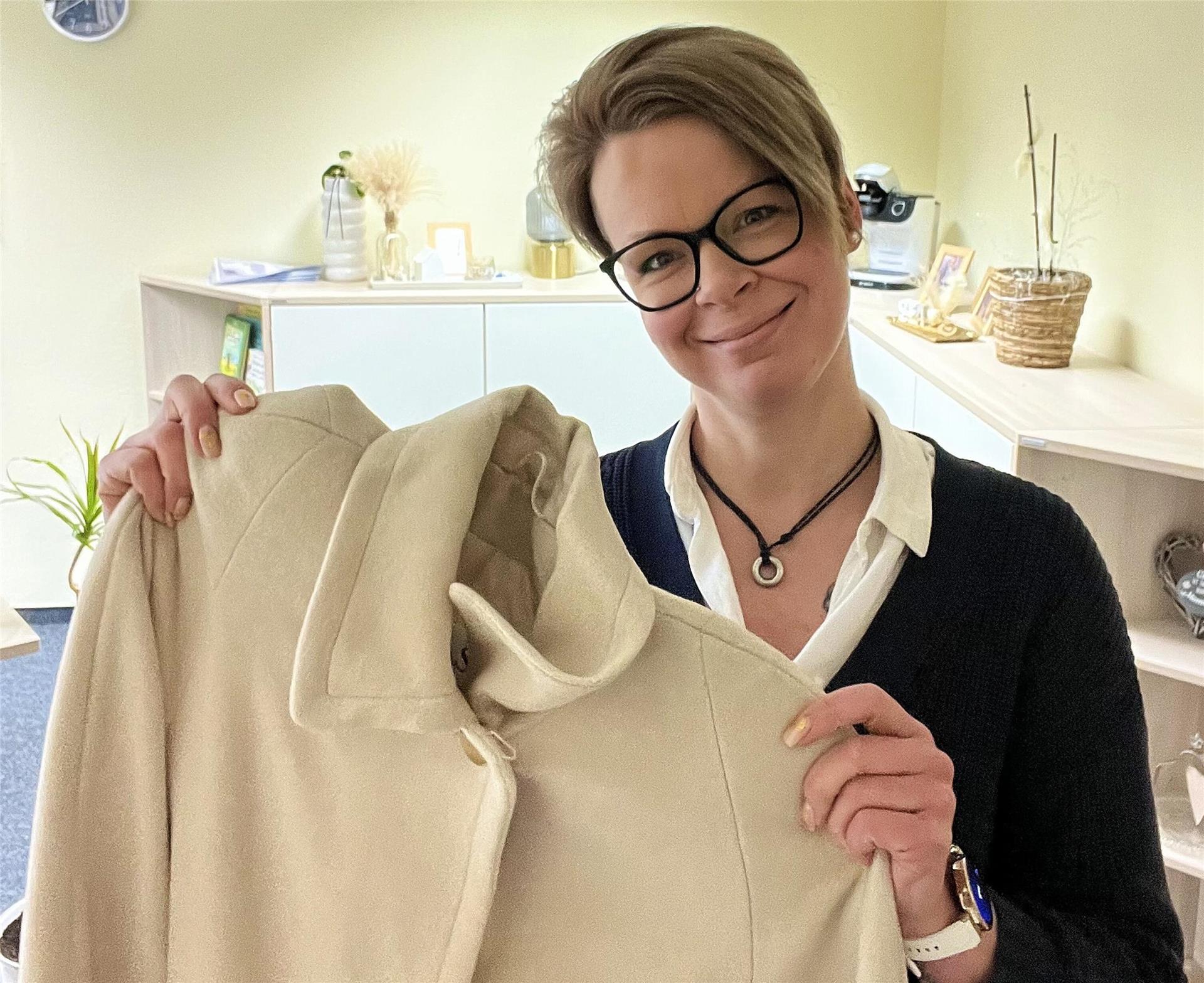 Eine lächelnde Frau mit Brille und kurzen Haaren hält einen beigen Mantel in die Kamera. Sie steht in einem freundlich gestalteten Büro mit Regalen, Pflanzen und Dekoration.
