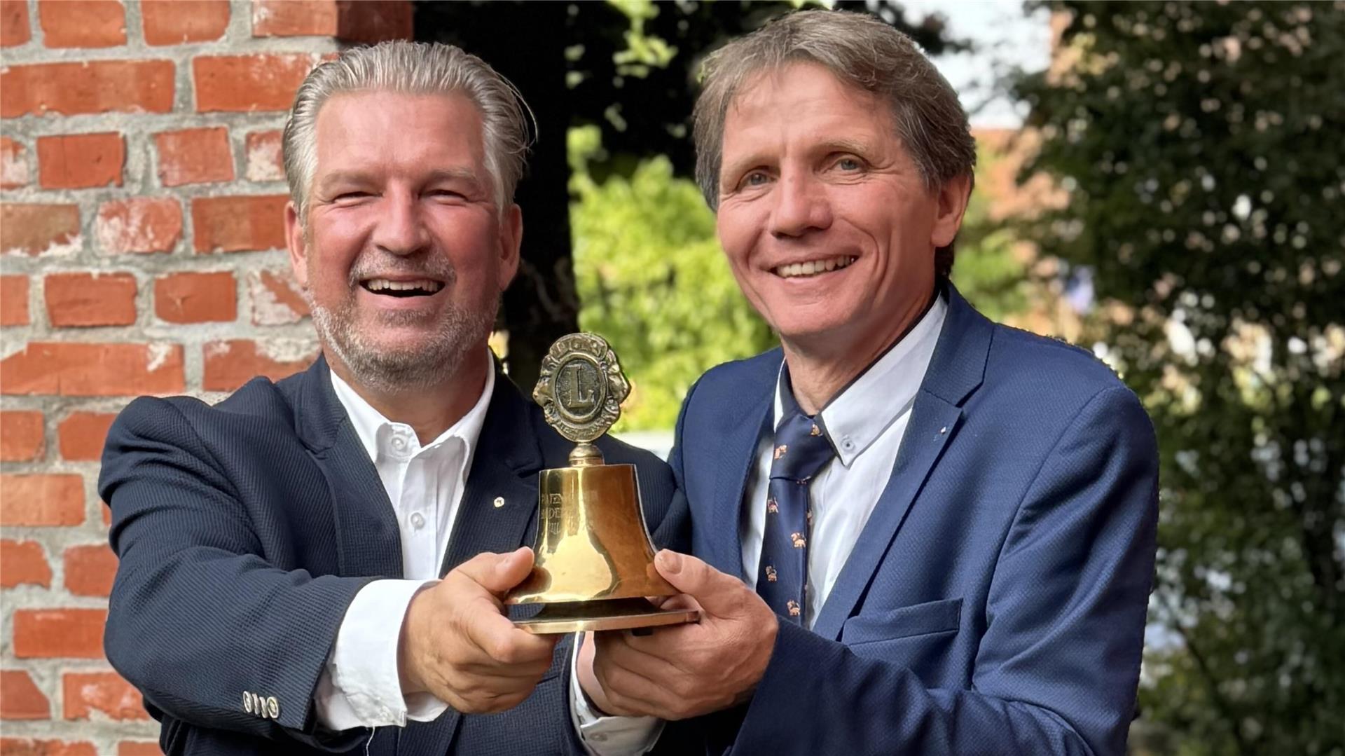 Jörg-Stephan Garms und Uwe Krüger stehen nebeneinander, lachen und halten gemeinsam eine goldene Lions-Club-Glocke, im Hintergrund eine Backsteinmauer und Bäume.