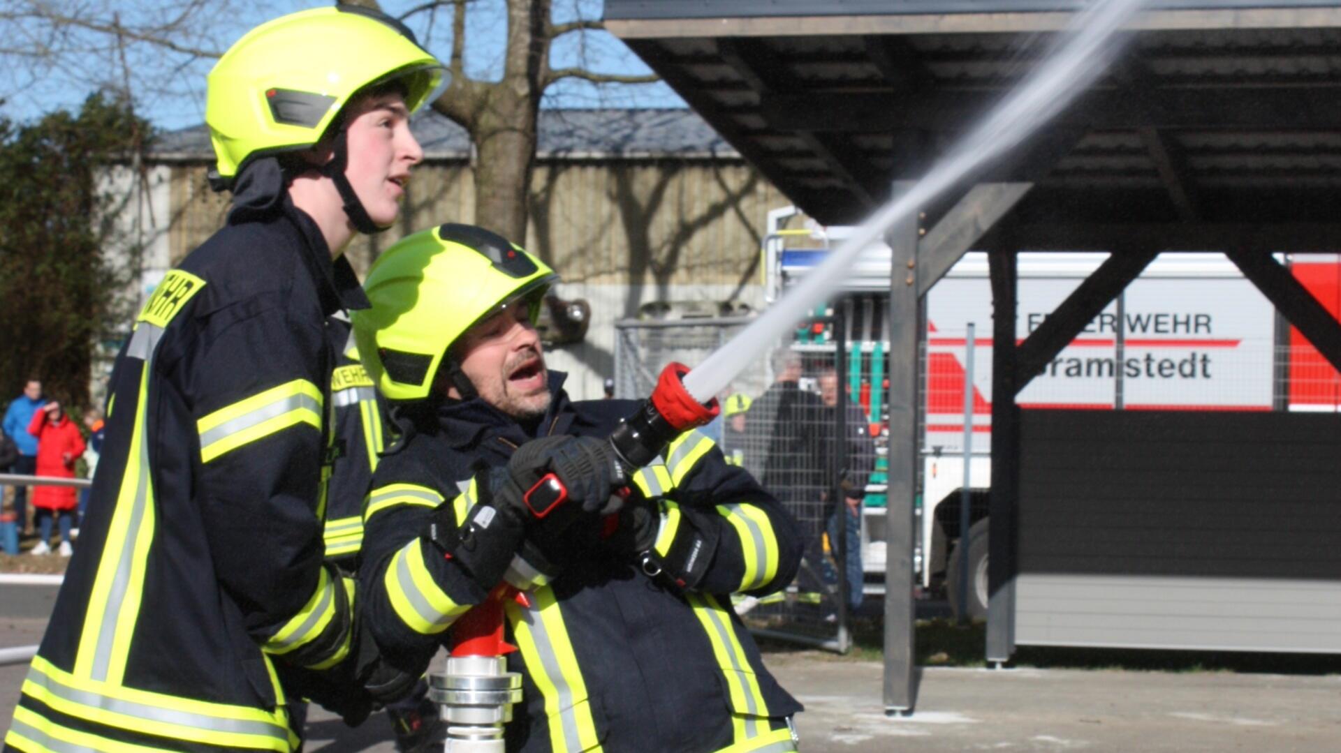 Zwei angehende Feuerwehrleute halten einen wassersprühenden Schlauch fest. Im Hintergrund steht ein Feuerwehrwagen der Bramstedter Feuerwehr. 