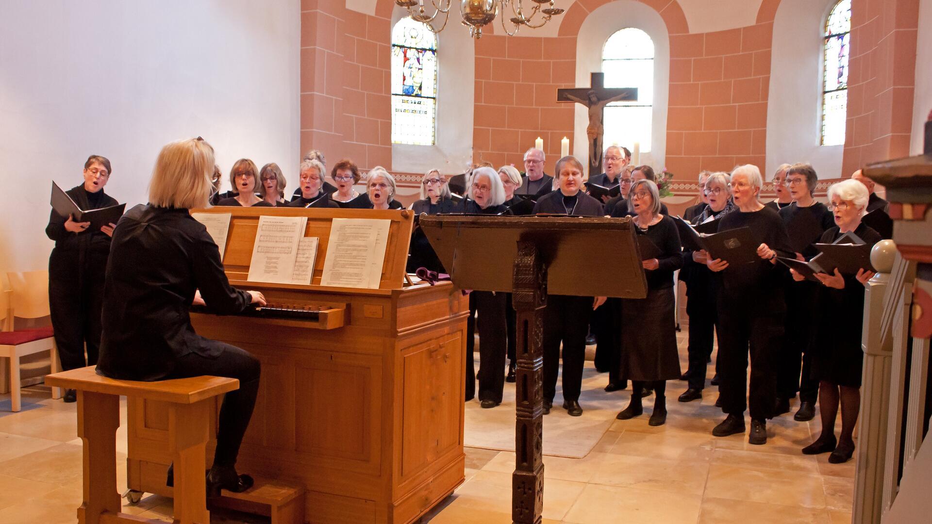 Eine Frau sitzt in einer Kirche an einer Orgel. Ihr steht ein Chor gegenüber und singt. 